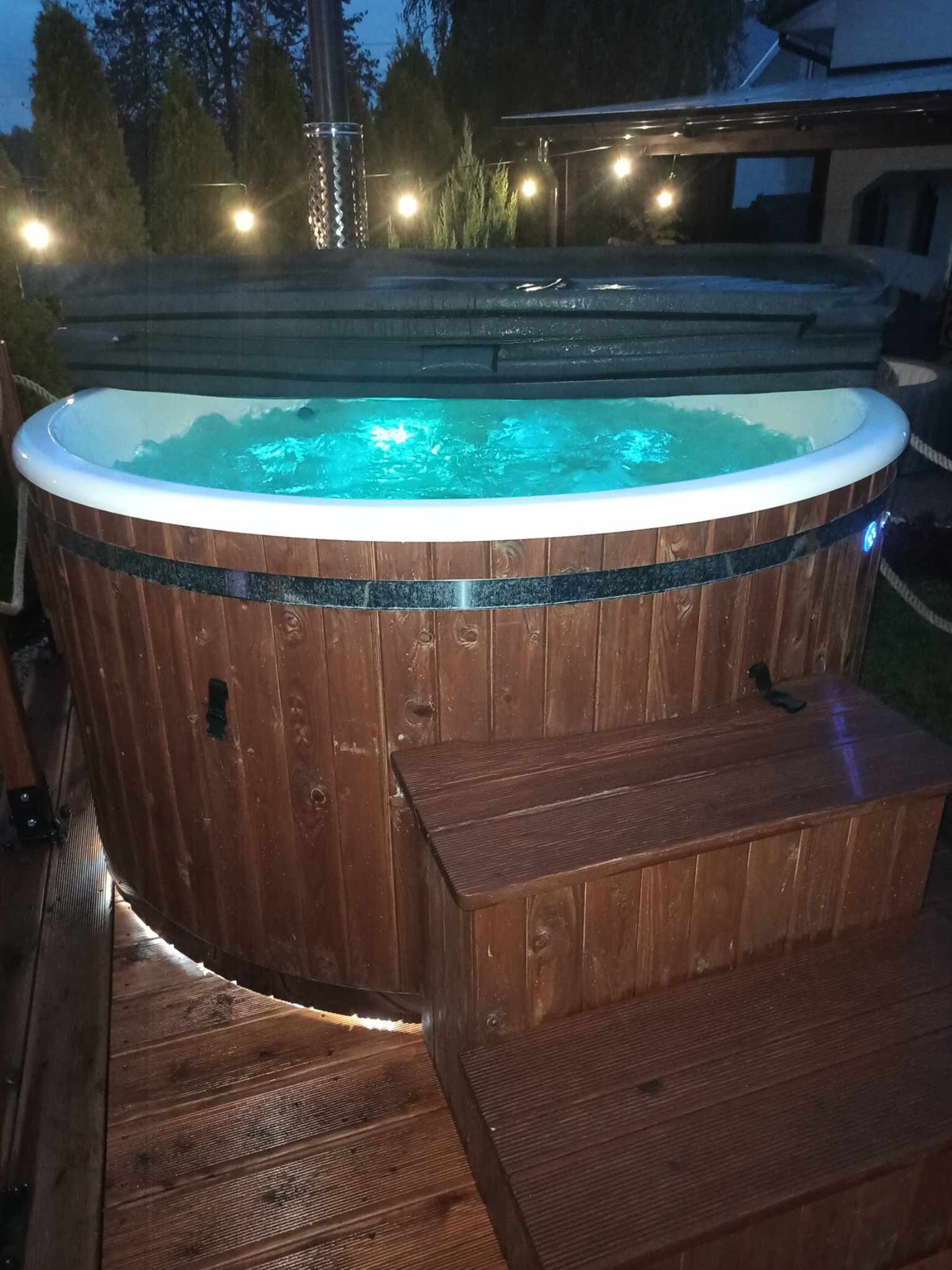 Drewniana balia ogrodowa z podświetloną wodą, z zewnątrz obłożona ciemnym drewnem, z pokrywą częściowo odsłoniętą, ustawiona na drewnianym tarasie z dwustopniowymi schodkami, w tle oświetlone...