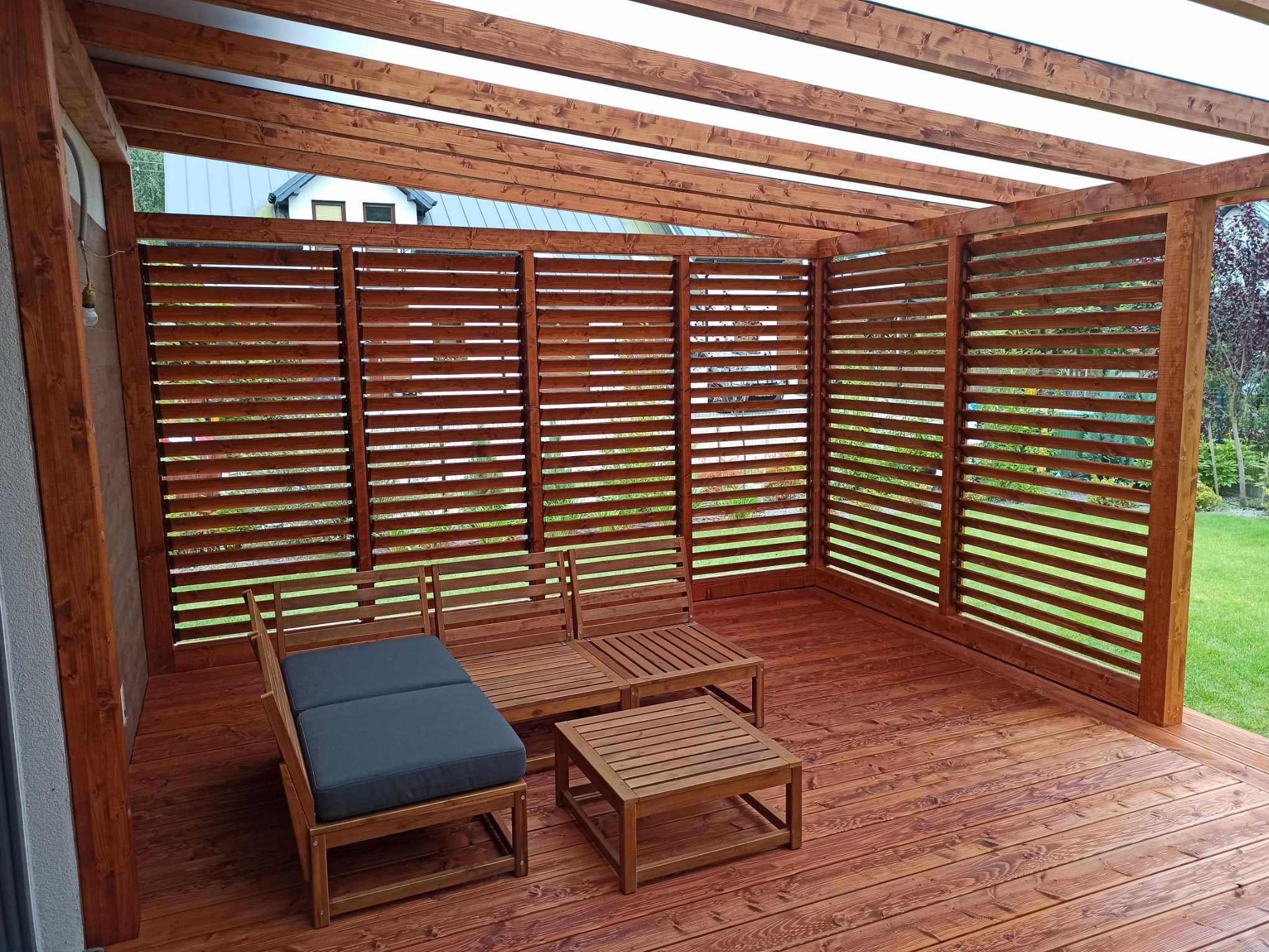 Zadaszony taras z drewnianą podłogą i lamelową ścianą, wyposażony w drewniane meble wypoczynkowe z szarymi poduszkami. Przez poziome szczeliny w ścianie widoczny zielony trawnik i drzewa w tle.