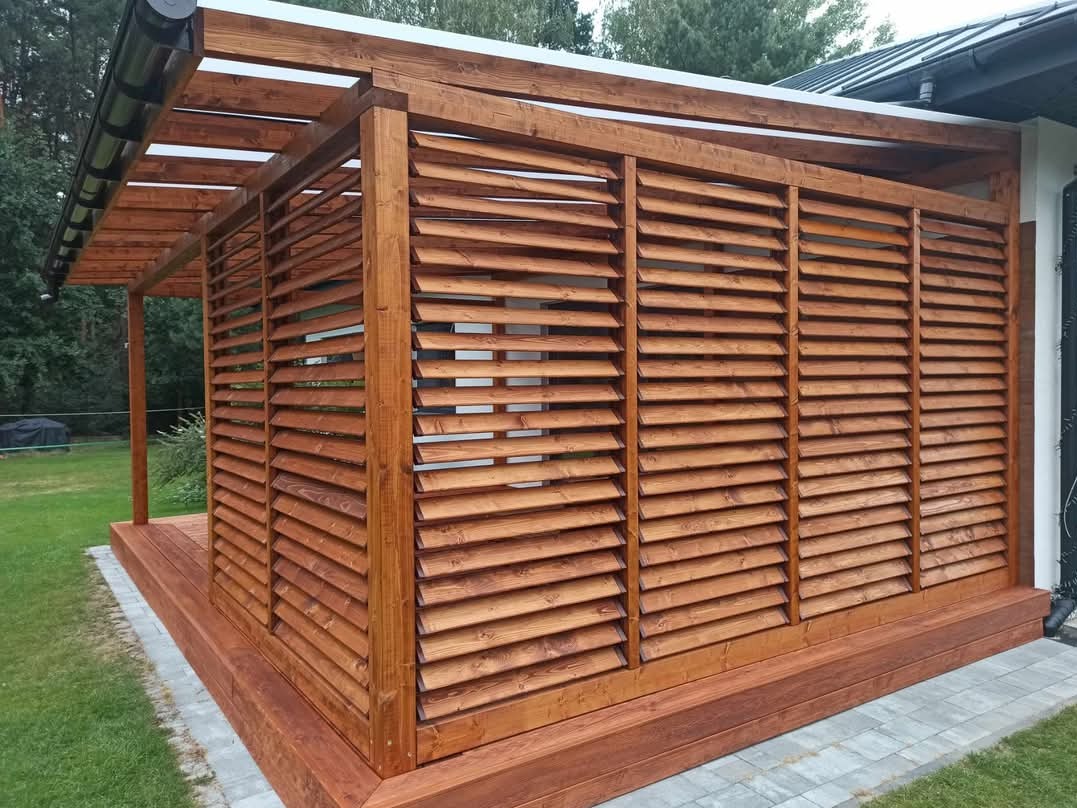 Zadaszona konstrukcja z drewnianymi żaluzjami, tworząca zacienione miejsce na tarasie, z widoczną konstrukcją dachu i elementami wykończenia z ciemnego drewna.