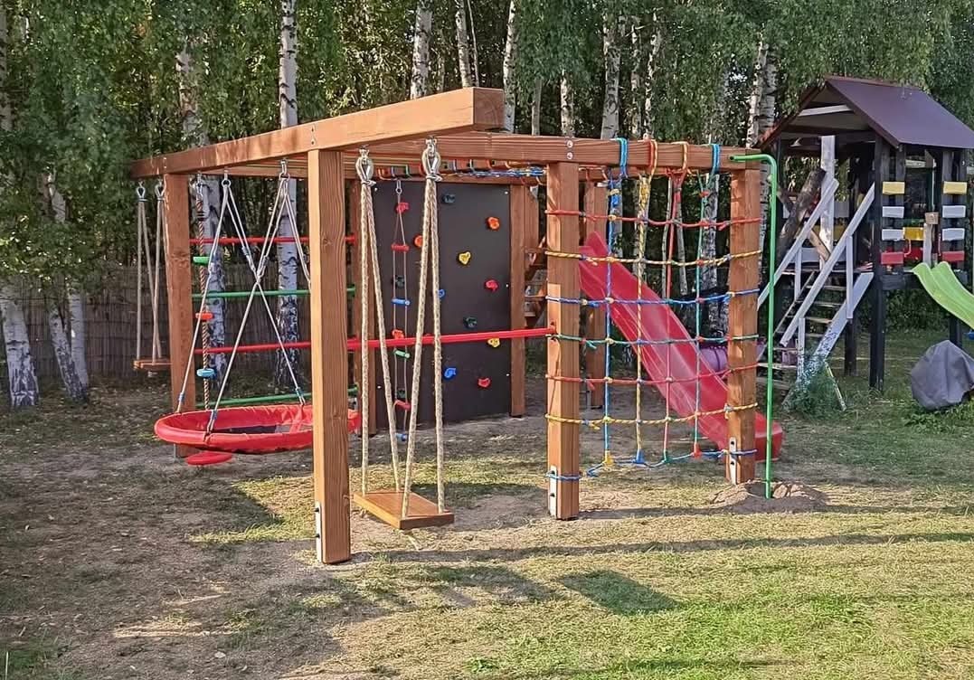Wielofunkcyjny plac zabaw z drewna: huśtawki, ścianka wspinaczkowa z kolorowymi uchwytami, siatka do wspinania i zjeżdżalnia. Dodatkowo widoczny domek na palach ze schodami i zjeżdżalnią w tle...