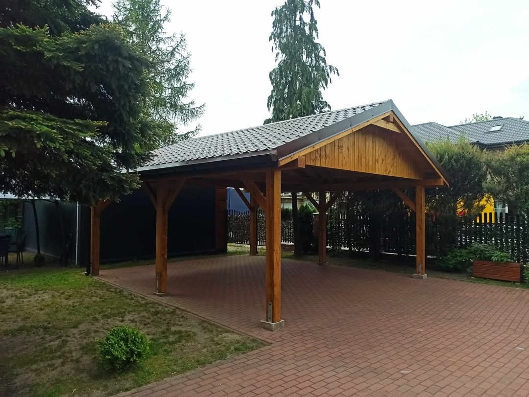 Drewniana konstrukcja z zadaszeniem z dachówki na ceglanym podłożu, widok z zewnątrz, otoczona zielenią i ogrodzeniem z metalowych prętów.