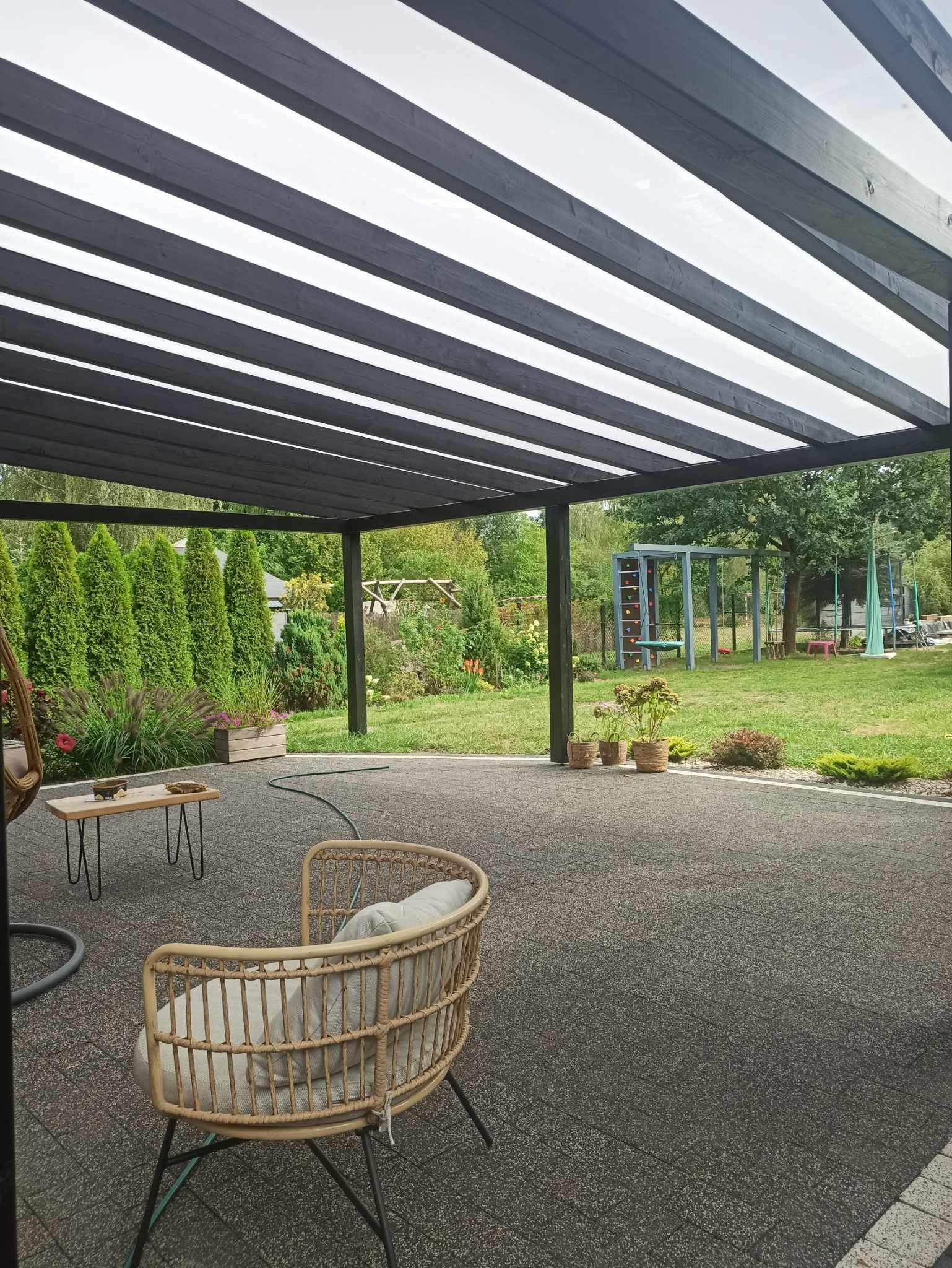 Nowoczesna pergola z czarnymi belkami i przezroczystym zadaszeniem, widok z dołu na taras z rattanowym fotelem i stolikiem kawowym, w tle zielony ogród z placem zabaw.