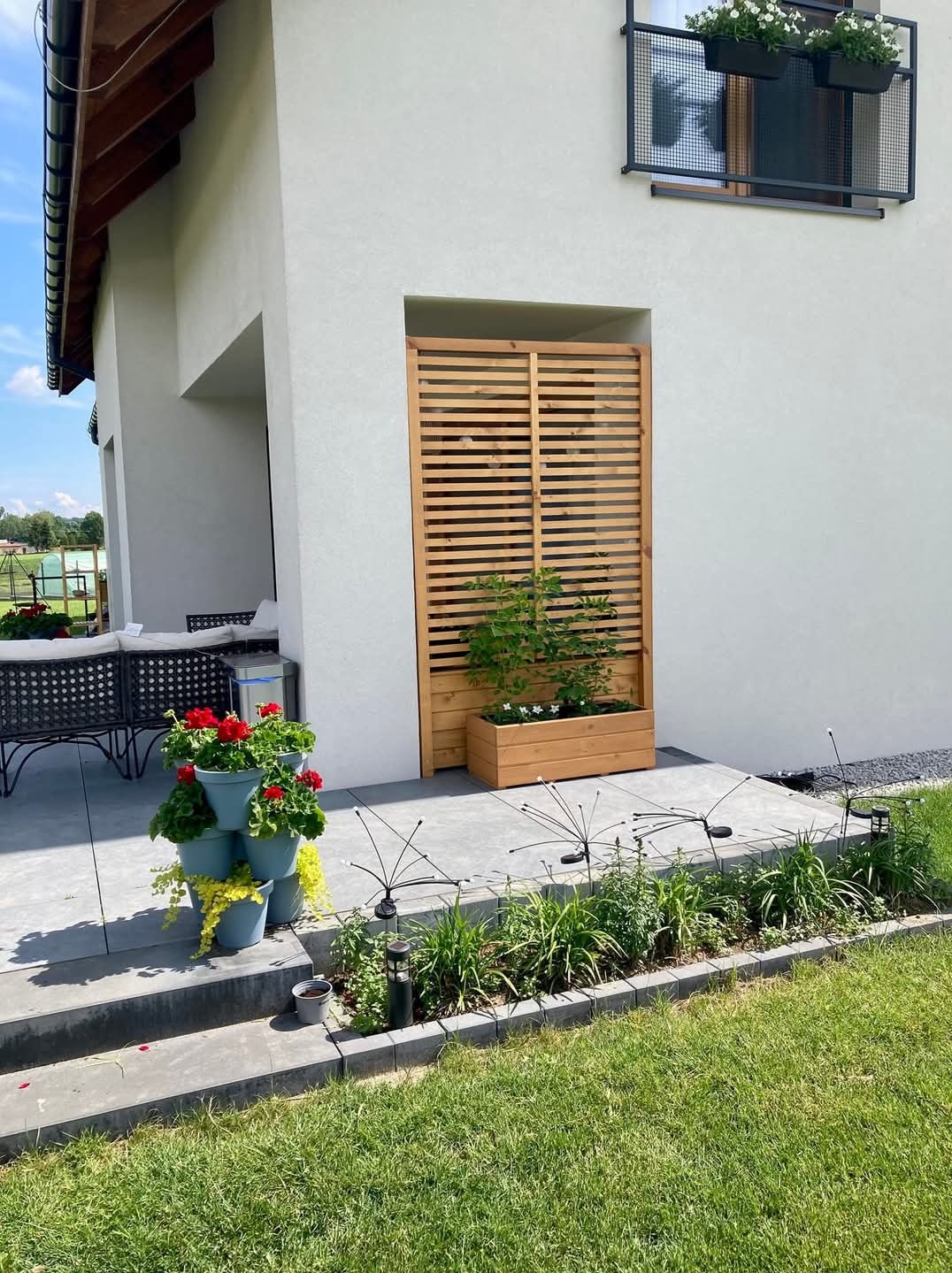 Nowoczesny taras z drewnianą przesłoną i donicą z roślinami, widok z poziomu trawnika, w tle biała ściana domu z czarnymi akcentami.