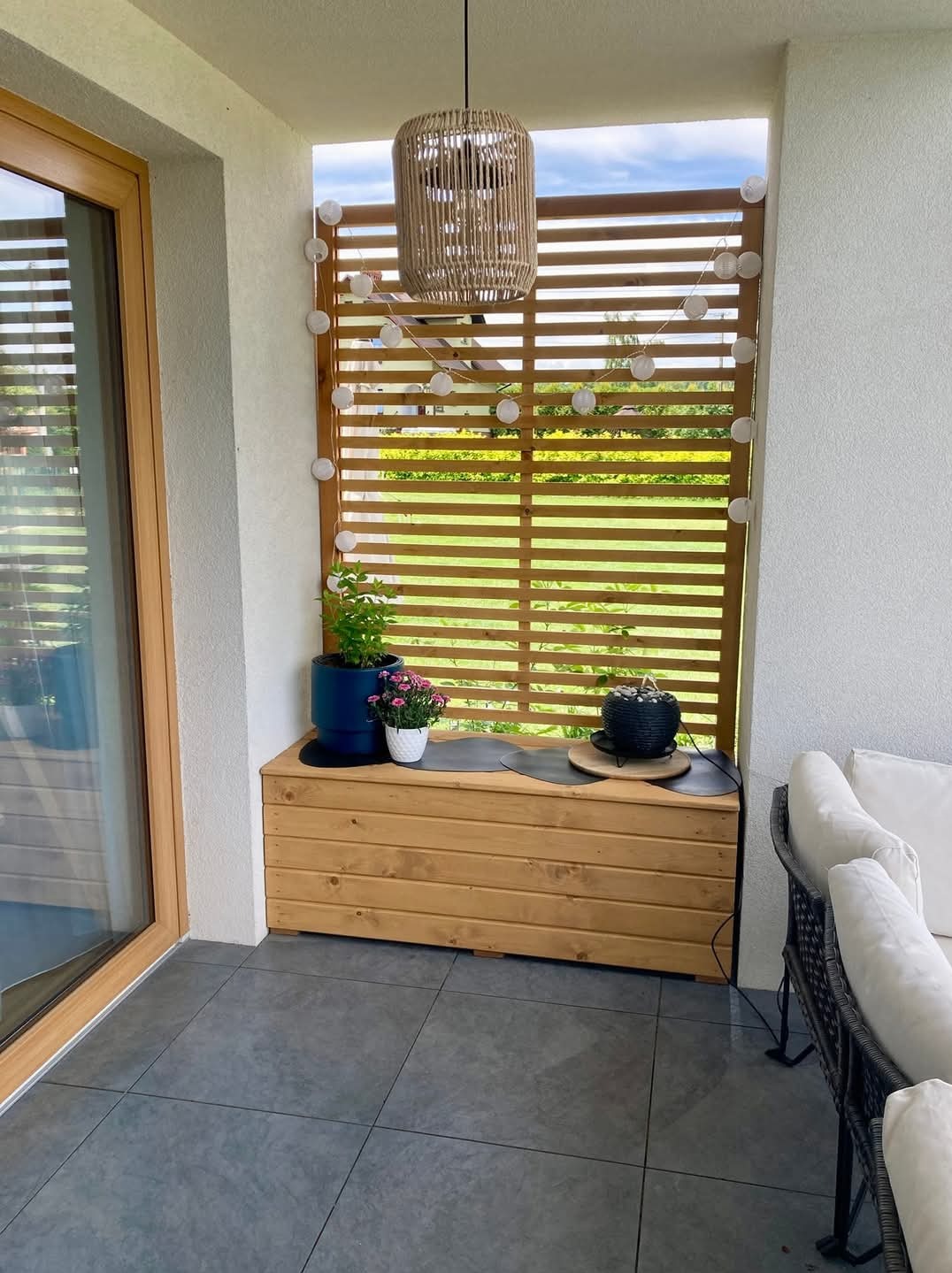 Zadaszony balkon z drewnianą ścianką ażurową ozdobioną girlandą świetlną, drewnianą skrzynią pełniącą funkcję siedziska i dekoracji w postaci doniczek z kwiatami oraz plecioną lampą wiszącą.