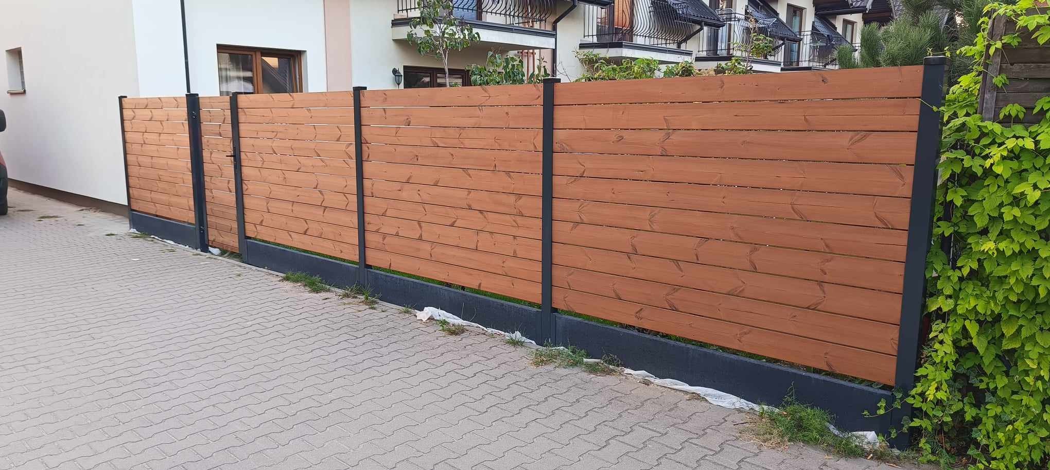 Nowoczesne ogrodzenie drewniane z czarnymi słupkami w Radziejowicach, widok z poziomu chodnika, z fragmentem zieleni po prawej stronie.