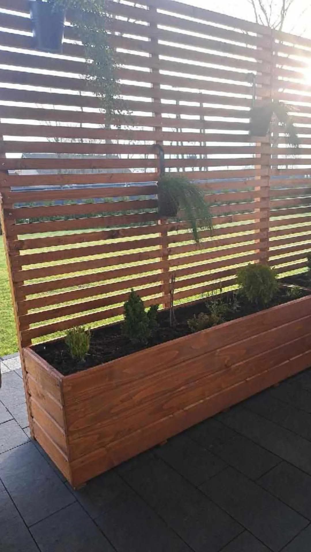 Drewniana konstrukcja z poziomych listew, pełniąca funkcję osłony i donicy, z posadzonymi roślinami, na tle trawnika w słoneczny dzień.