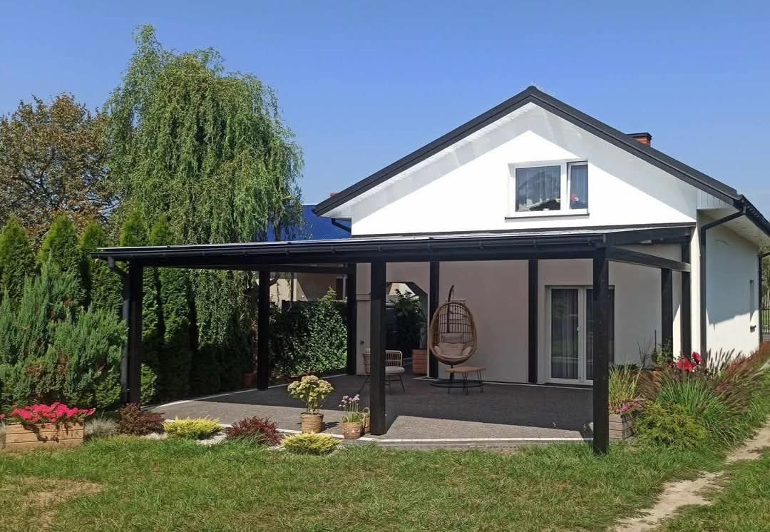 Zadaszony taras przydomowy z czarnymi słupami i belkami, wyposażony w wiszące krzesło i meble ogrodowe, otoczony zielenią i kwiatami w donicach, na tle białego domu z czarnym dachem.