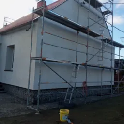 Dom jednorodzinny z szarą elewacją w trakcie prac, widoczne rusztowania, wiadro z farbą i szpachelka obok, cokół wykończony kamieniem.
