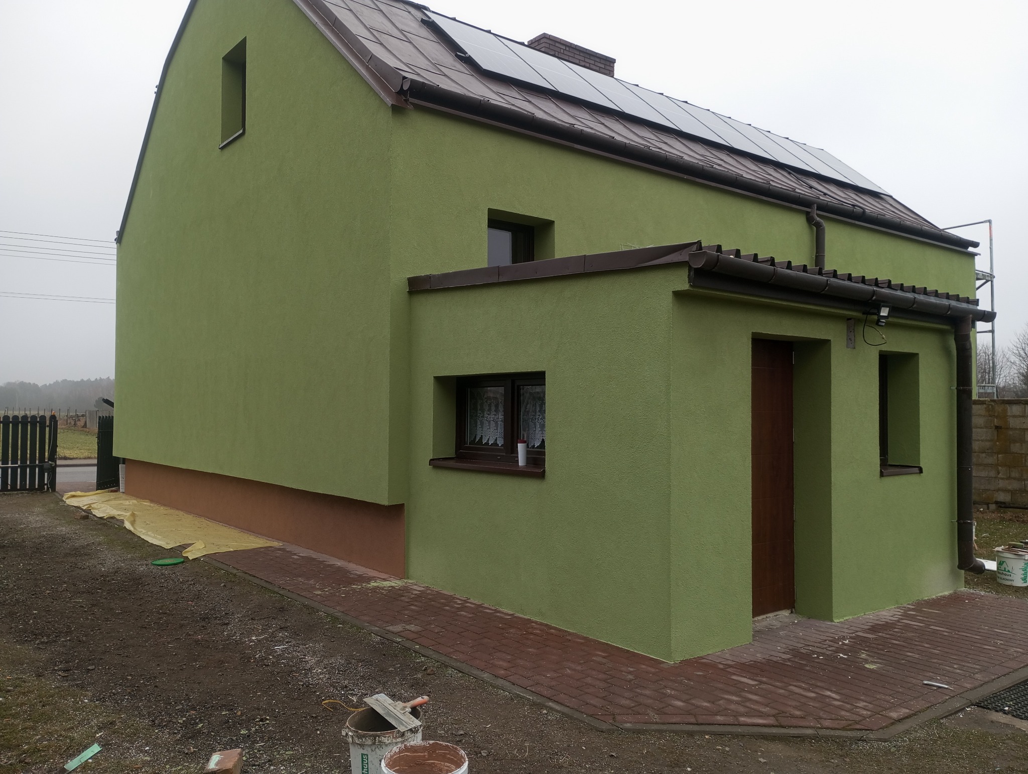 Dwukondygnacyjny dom z zieloną elewacją i brązowymi akcentami, widoczne panele słoneczne na dachu, teren wokół domu częściowo utwardzony kostką, widoczne narzędzia i farby malarskie.