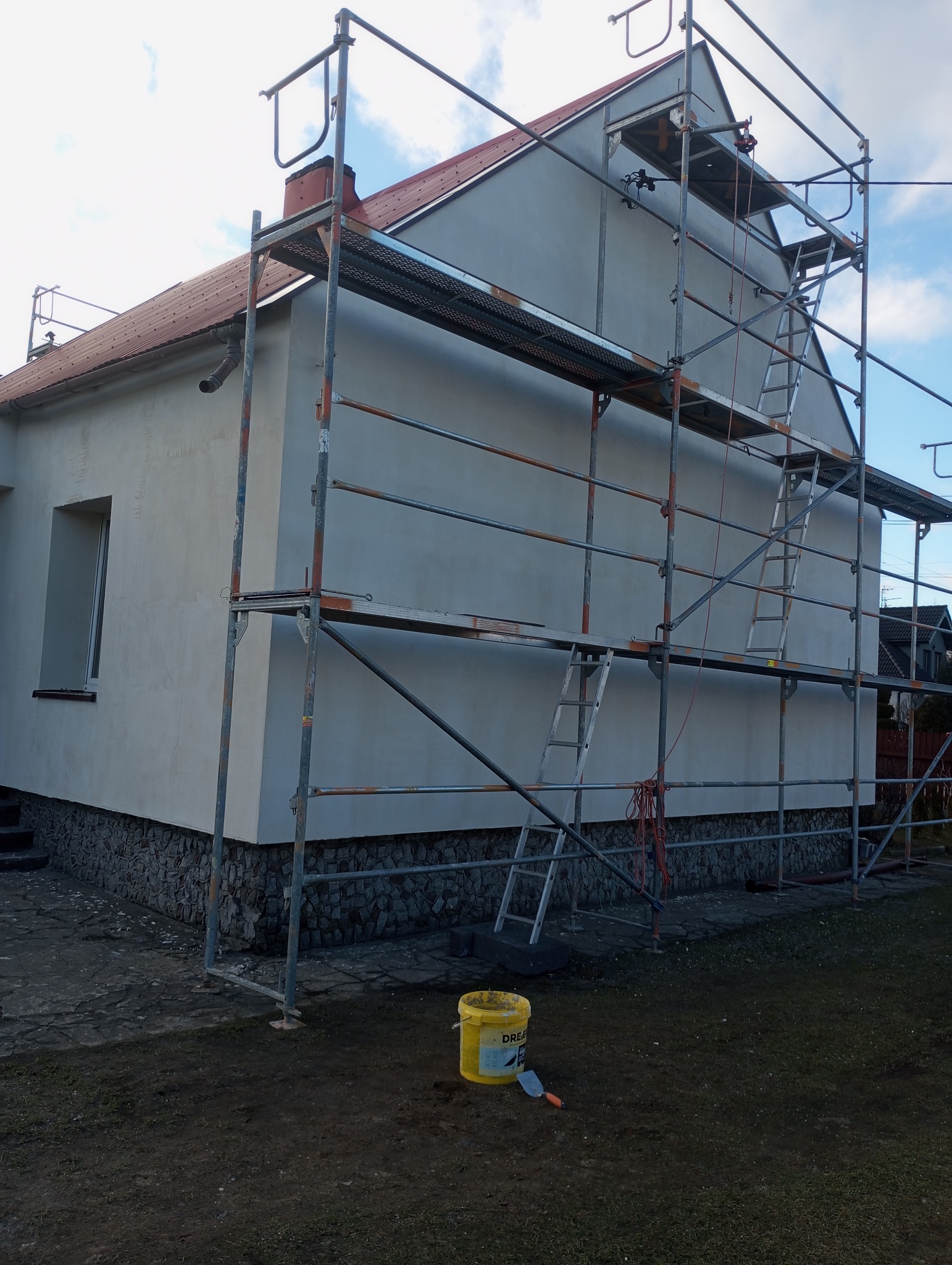 Dom jednorodzinny z szarą elewacją w trakcie prac, widoczne rusztowania, wiadro z farbą i szpachelka obok, cokół wykończony kamieniem.