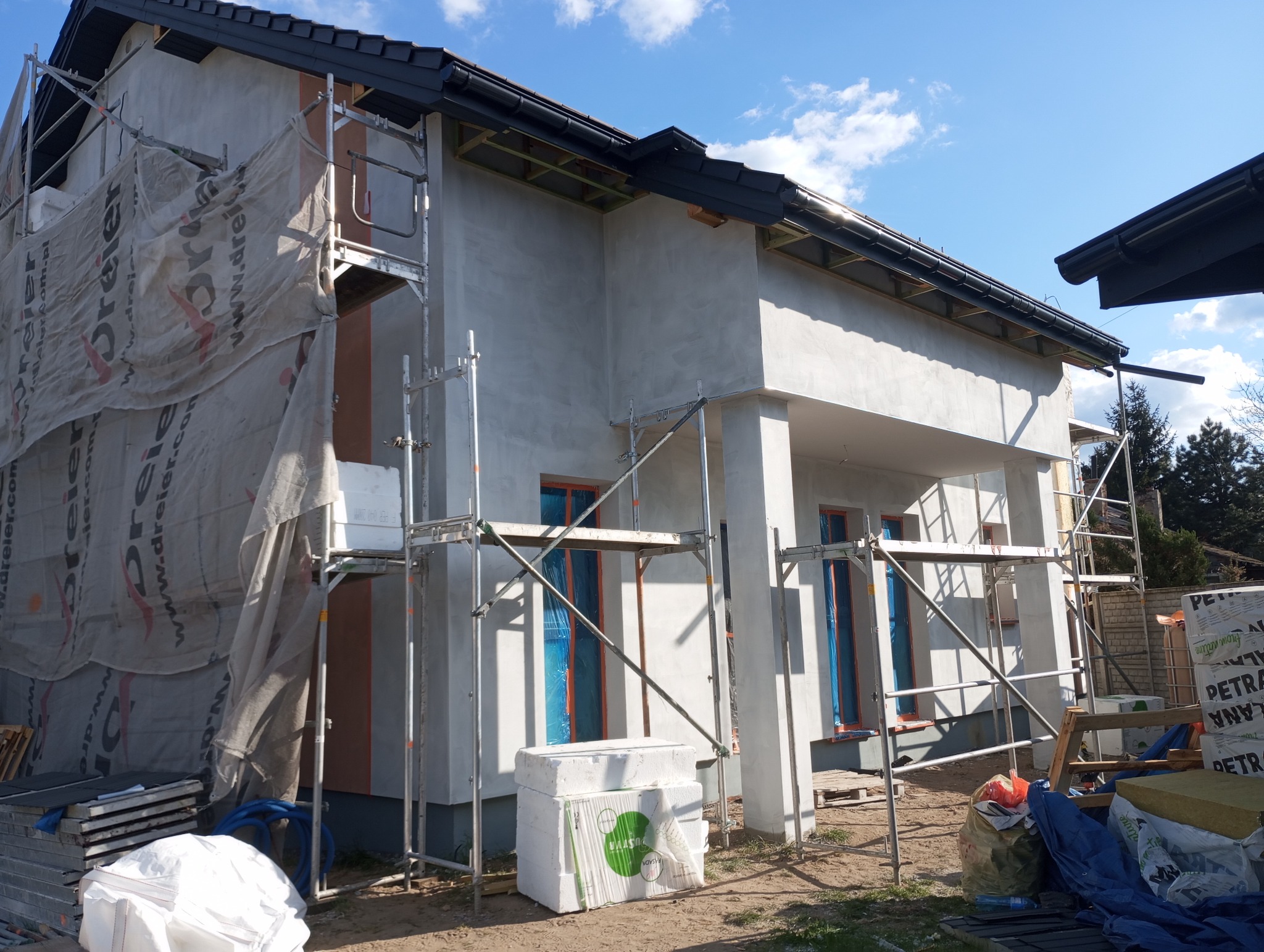 Dom w trakcie ocieplania: styropian, rusztowania, zabezpieczone okna folią. Częściowo otynkowana fasada w jasnym odcieniu. Widoczne materiały budowlane.