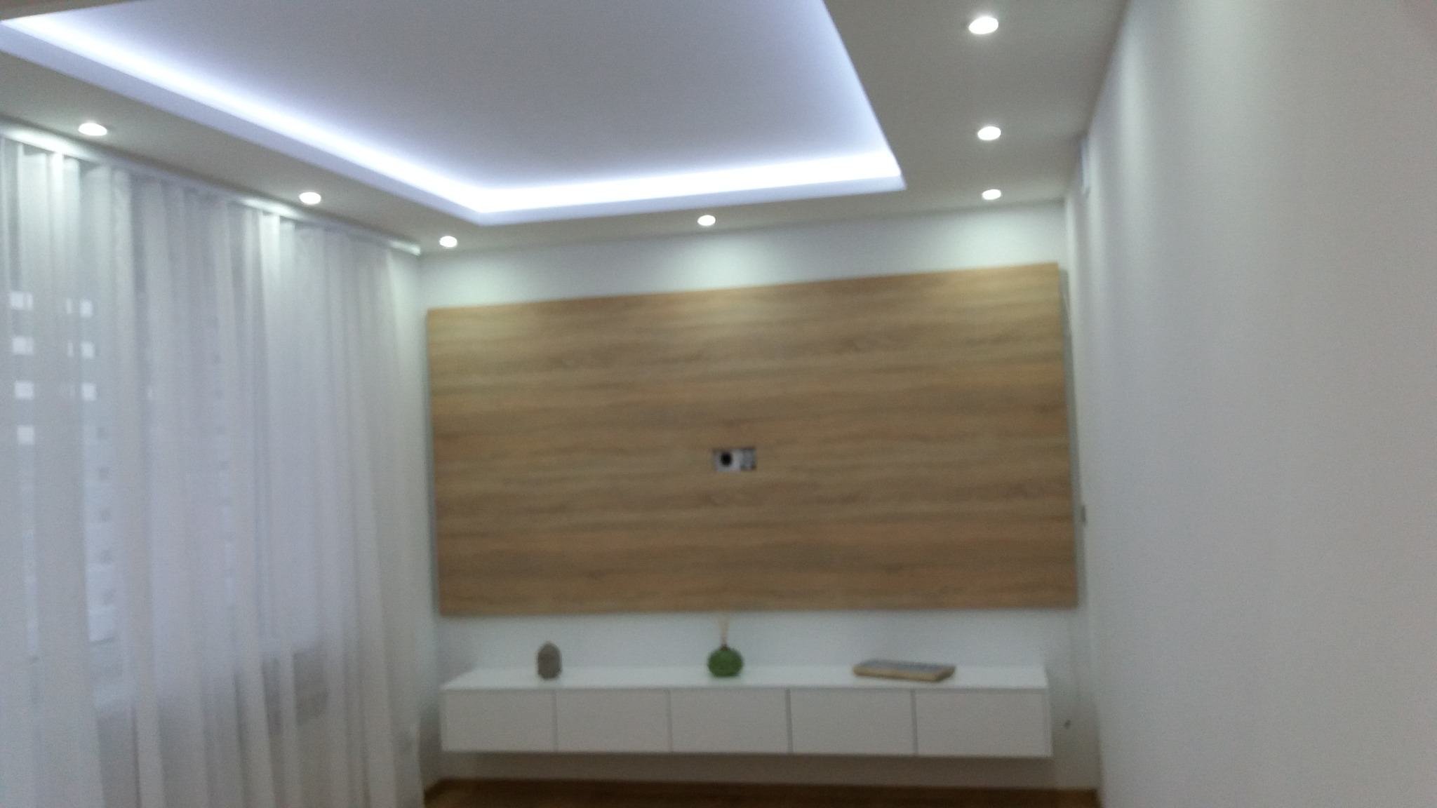 Jasne wnętrze pokoju z drewnianą ścianą dekoracyjną, białymi zasłonami i podwieszanym sufitem z oświetleniem punktowym i LED, minimalistyczna biała szafka RTV z dekoracjami.