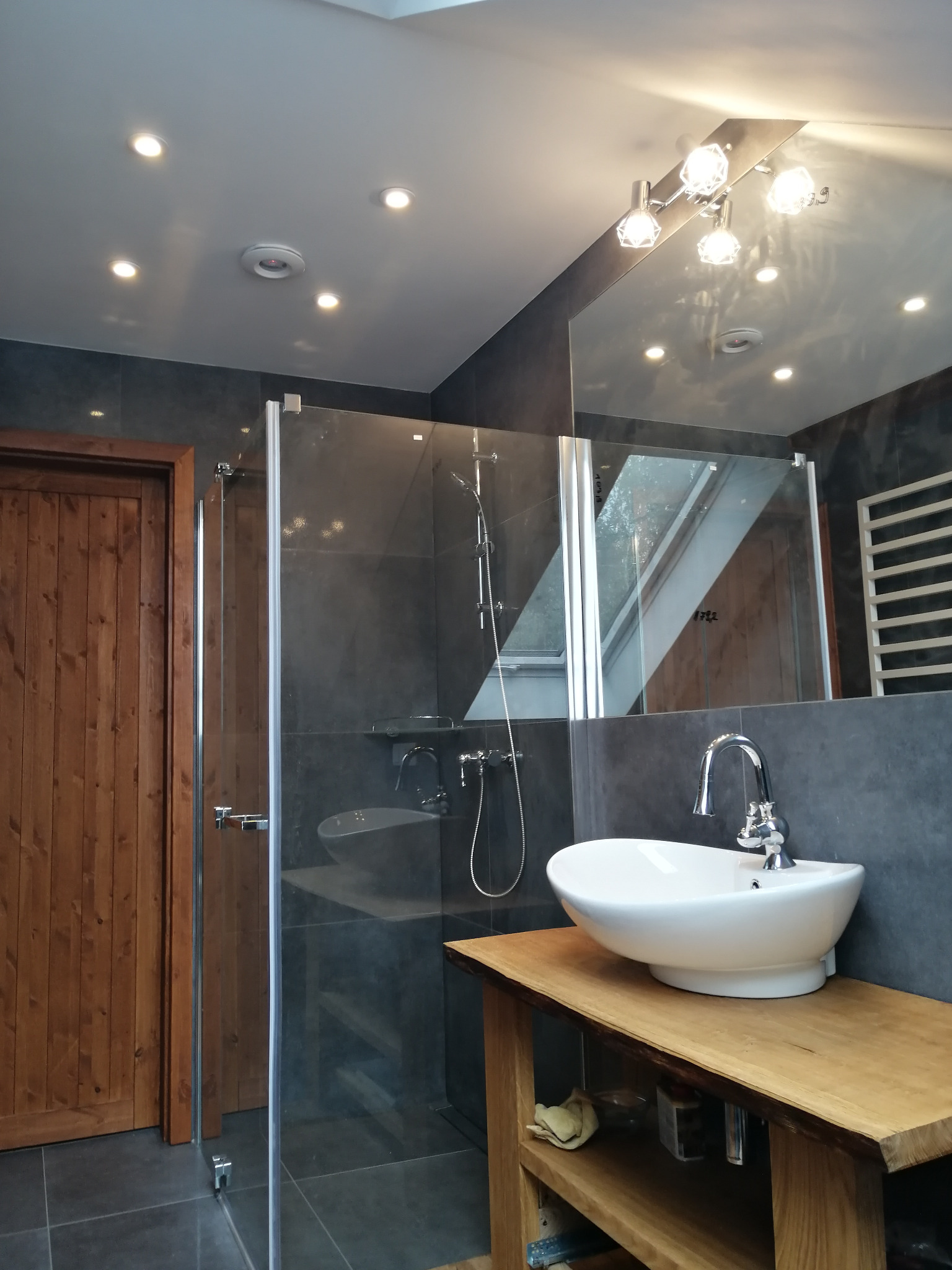 Nowoczesna łazienka z kabiną prysznicową ze szklanymi drzwiami, umywalką nablatową na drewnianym blacie z półką, szarą glazurą na ścianach i suficie z oświetleniem punktowym.