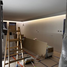 PREMIUM EKSPERT - Wnętrze w trakcie prac wykończeniowych: drabina malarska, wałek i kuweta na podłodze zabezpieczonej folią, ściany przygotowane do malowania z oświetleniem LED.