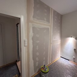 PREMIUM EKSPERT - Ściana po montażu płyt karton-gips i wstępnym szpachlowaniu, widoczny kubeł z narzędziami, zabezpieczona podłoga, fragment futryny z włącznikiem.