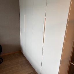 PREMIUM EKSPERT - Biała, minimalistyczna szafa wnękowa z podłużnymi uchwytami, na tle szarej ściany i drewnianej podłogi, widziana pod kątem z boku.