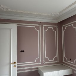 PREMIUM EKSPERT - Eleganckie wykończenie wnętrza: pastelowa ściana z białymi listwami dekoracyjnymi, sztukateria sufitowa, białe drzwi i wisząca półka. Widoczne gniazdko elektryczne.
