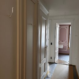 PREMIUM EKSPERT - Korytarz w trakcie wykańczania: białe ściany z listwami, zabezpieczona podłoga, widoczne drzwi do pokoju z dekoracyjną ścianą w odcieniach różu i bieli.