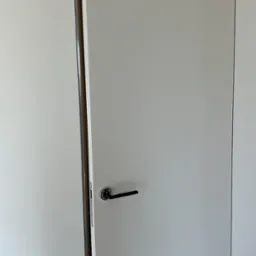 Minimalistyczne, białe drzwi wewnętrzne uchylone, z czarną klamką na tle jasnej ściany. Widoczna ościeżnica i fragment pomieszczenia za drzwiami.