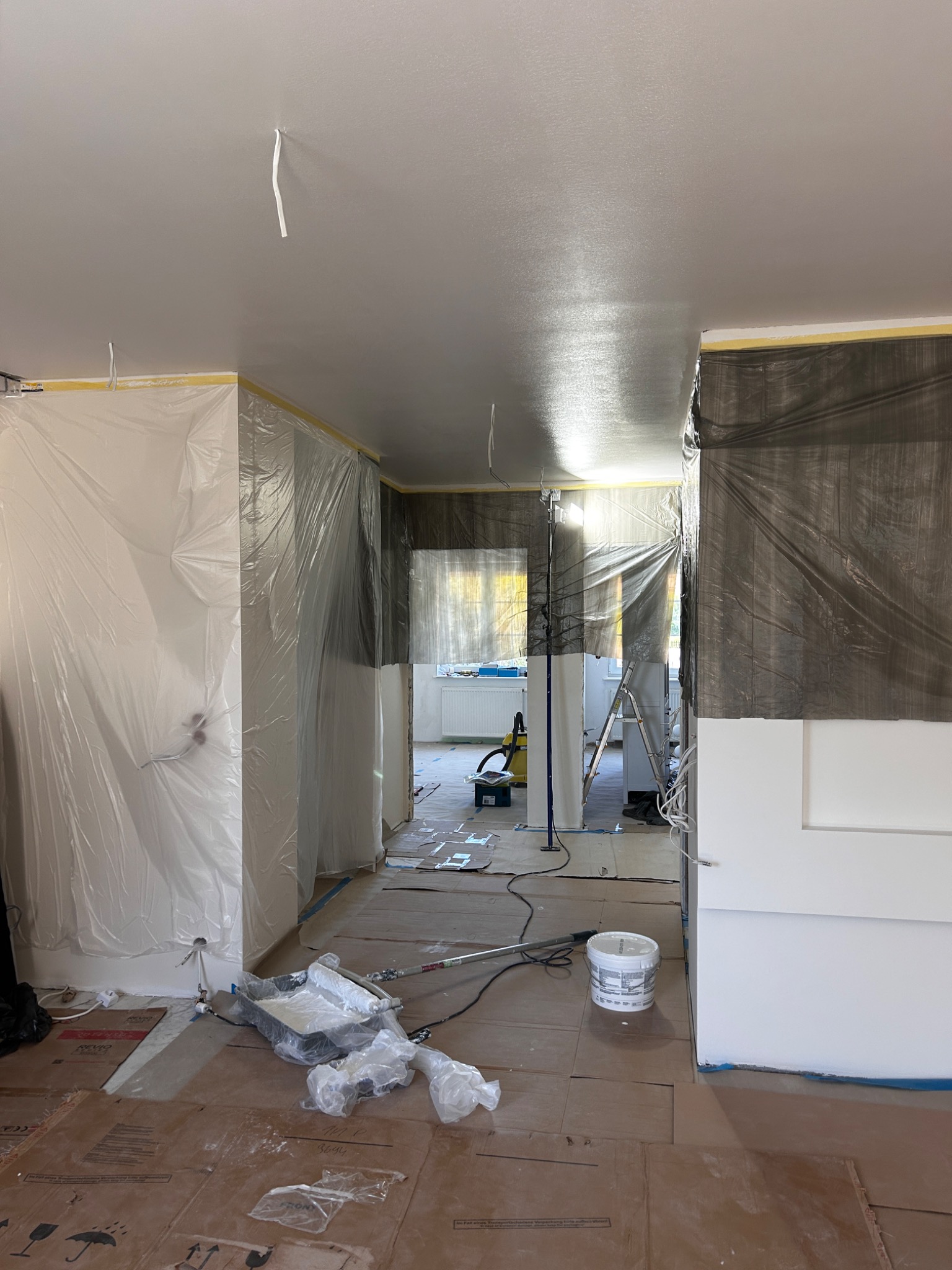 Wnętrze w trakcie malowania: świeżo pomalowany sufit, zabezpieczone ściany folią i taśmą, widoczny sprzęt malarski i wiadro z farbą. Przygotowanie do malowania ścian.