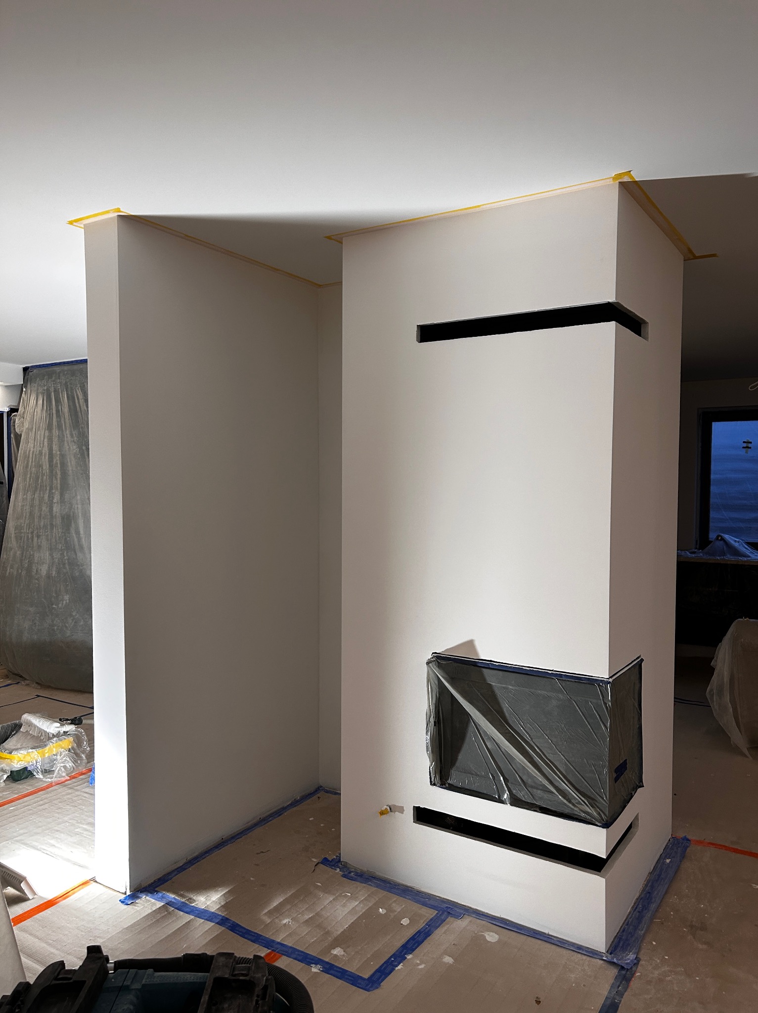 Wnętrze w trakcie prac wykończeniowych: świeżo pomalowane ściany z maskowaniem taśmą, zabezpieczony kominek folią, podłoga zakryta folią malarską.