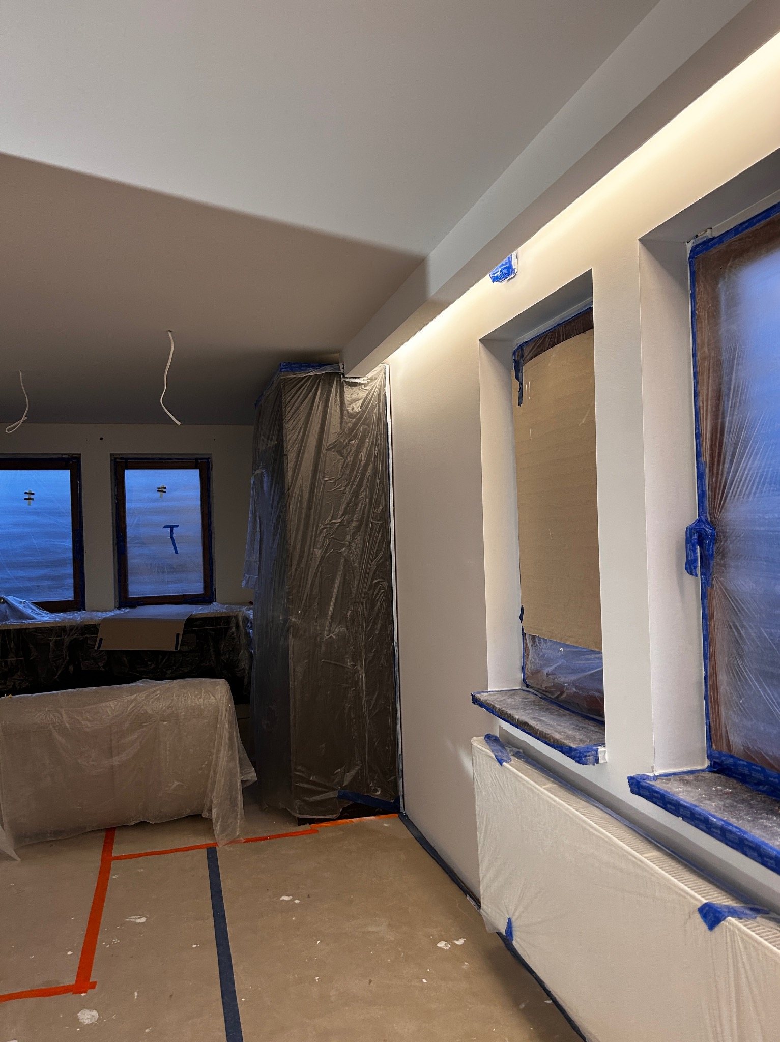 Wnętrze w trakcie odświeżania: świeżo pomalowane ściany w jasnym odcieniu, okna i meble zabezpieczone folią malarską, widoczne taśmy malarskie na podłodze.