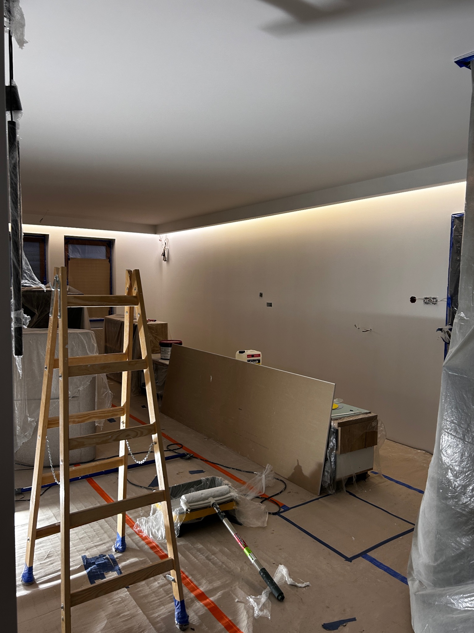 Wnętrze w trakcie prac wykończeniowych: drabina malarska, wałek i kuweta na podłodze zabezpieczonej folią, ściany przygotowane do malowania z oświetleniem LED.