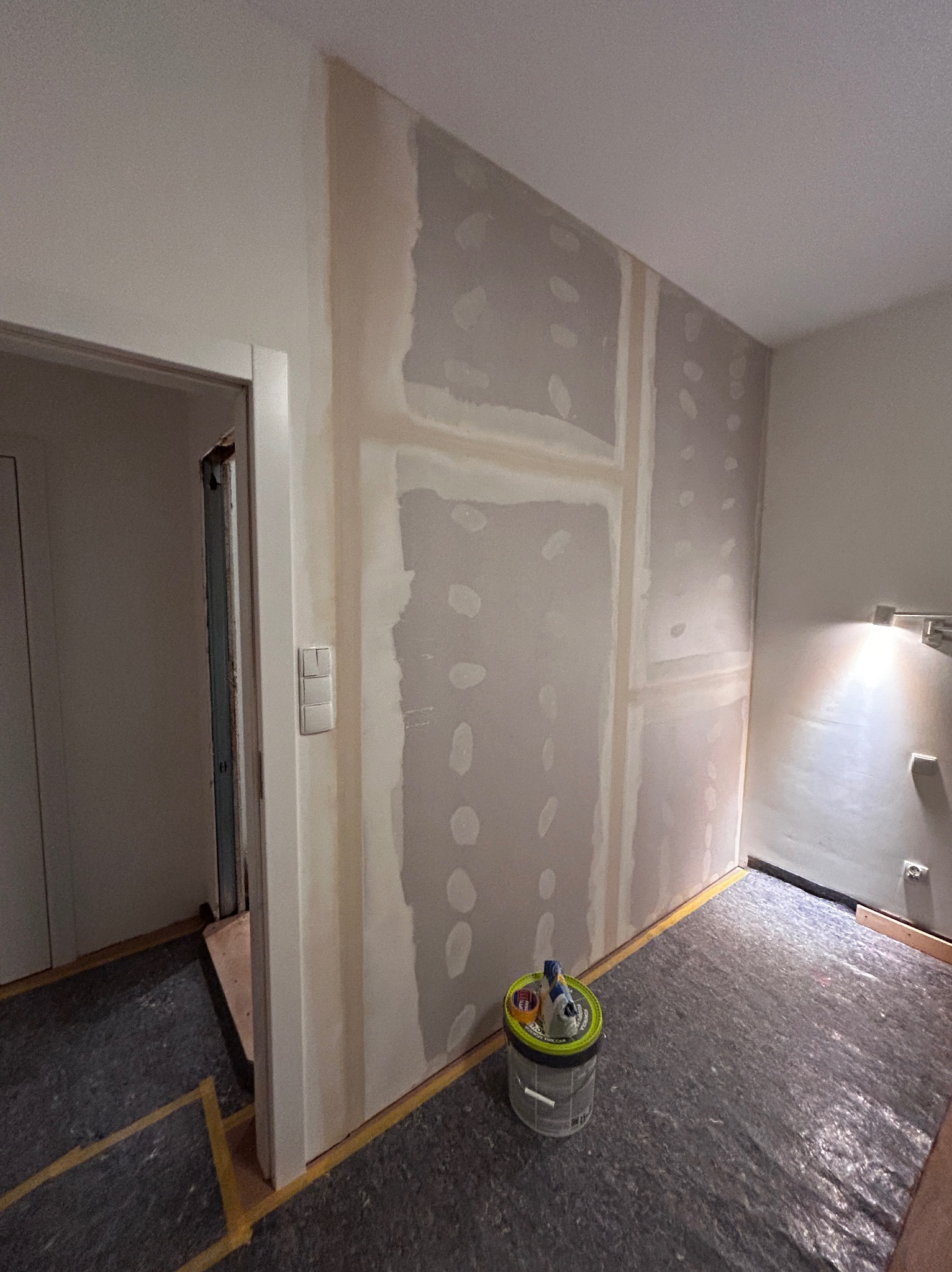 Ściana po montażu płyt karton-gips i wstępnym szpachlowaniu, widoczny kubeł z narzędziami, zabezpieczona podłoga, fragment futryny z włącznikiem.