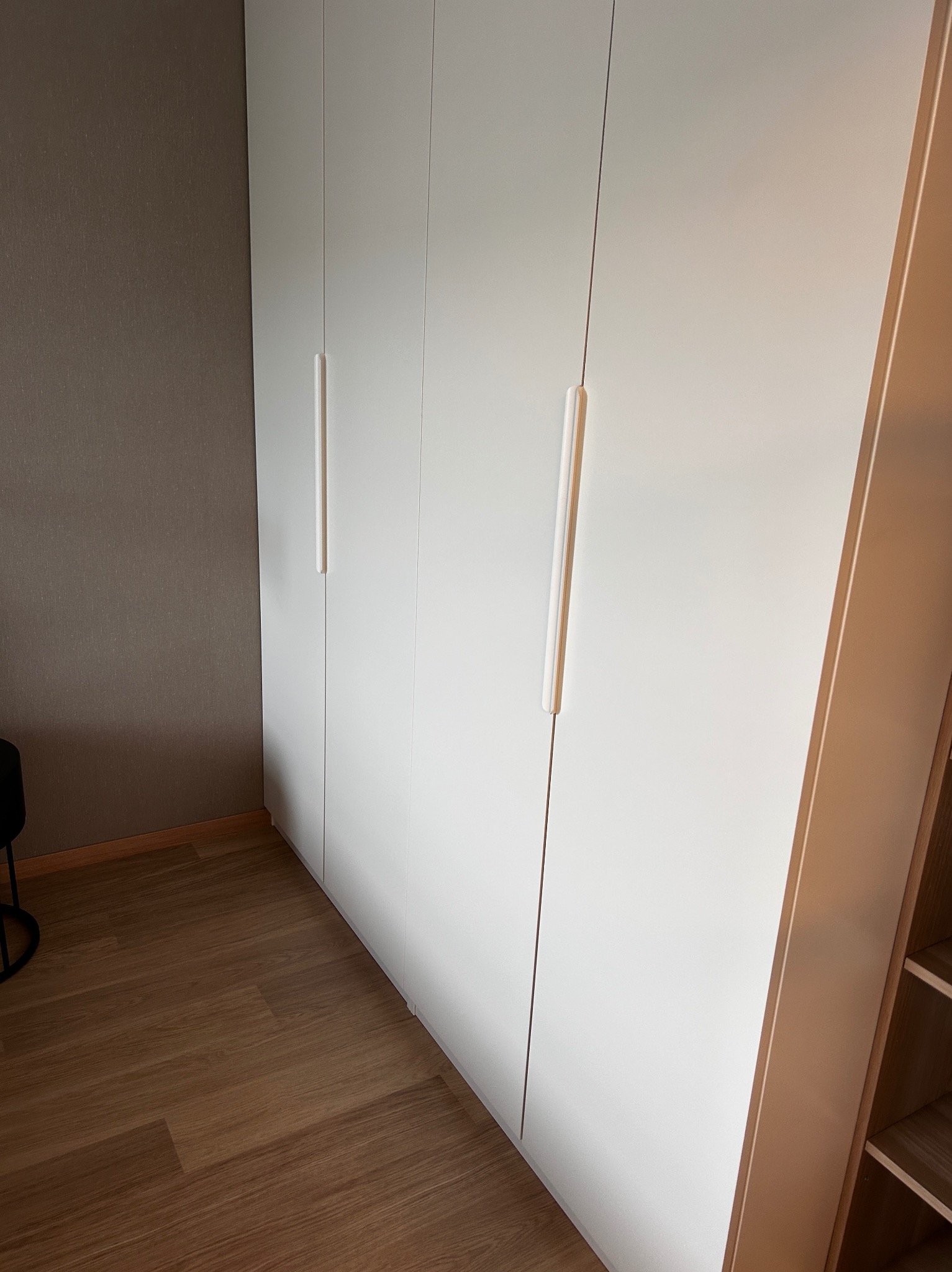 Biała, minimalistyczna szafa wnękowa z podłużnymi uchwytami, na tle szarej ściany i drewnianej podłogi, widziana pod kątem z boku.