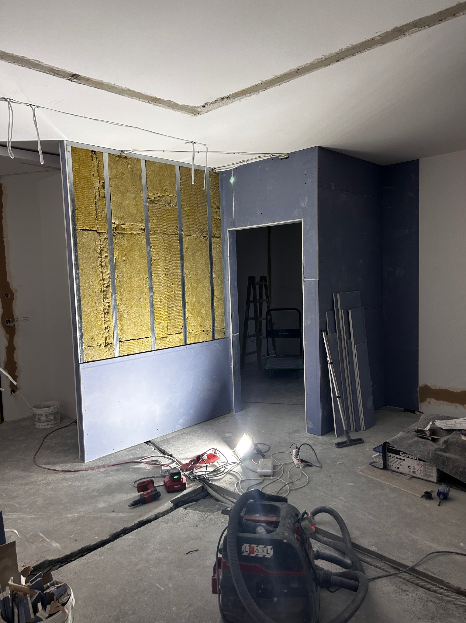 Wnętrze w trakcie prac: ściana z płyt karton-gips z widoczną izolacją, narzędzia na podłodze, sufit z wyciętymi elementami. Widoczny etap prac wykończeniowych.