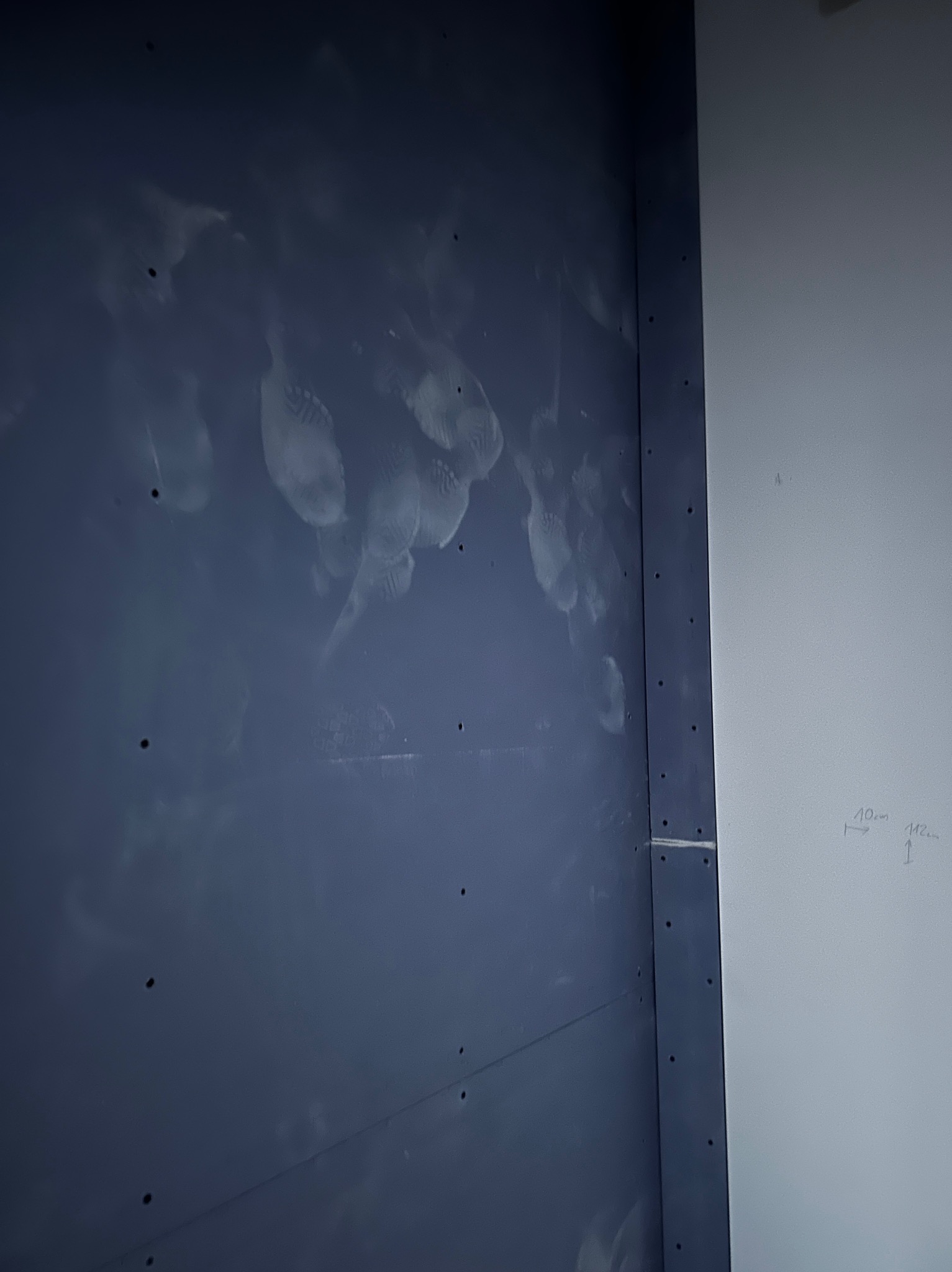 Narożnik pokoju: ściana pomalowana na granatowo ze śladami szpachli, sąsiadująca z białą ścianą z odręcznymi notatkami. Widoczne otwory po wkrętach.