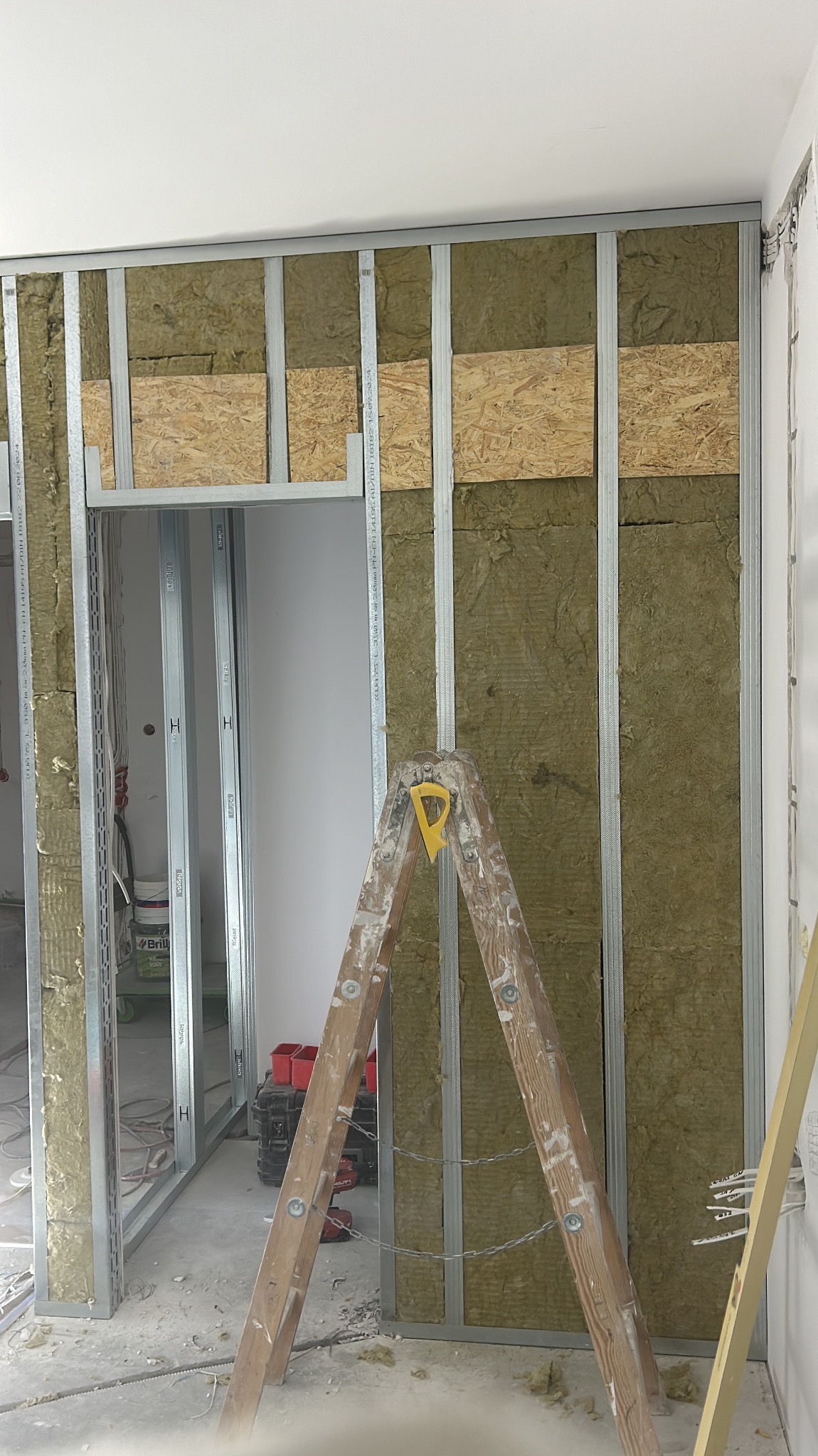 Montaż ścianki działowej z metalowych profili, wypełnionej izolacją i płytami OSB. Widoczna drewniana drabina i narzędzia. Etap prac wykończeniowych.