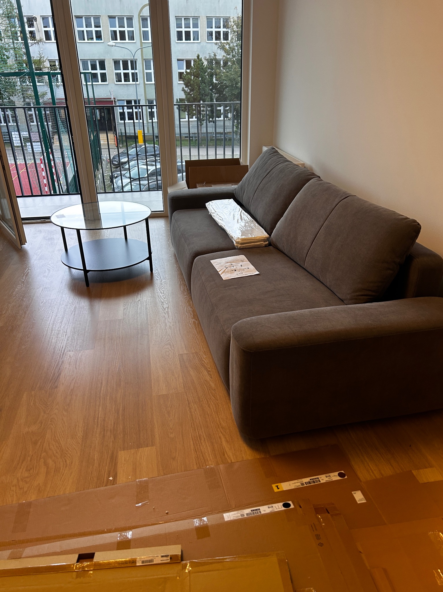 Nowoczesny salon z drewnianą podłogą, szarą sofą z poduszkami i okrągłym stolikiem kawowym. Widok z balkonu na budynek szkolny.