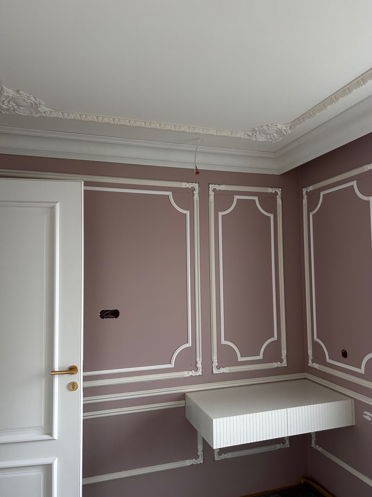 Eleganckie wykończenie wnętrza: pastelowa ściana z białymi listwami dekoracyjnymi, sztukateria sufitowa, białe drzwi i wisząca półka. Widoczne gniazdko elektryczne.