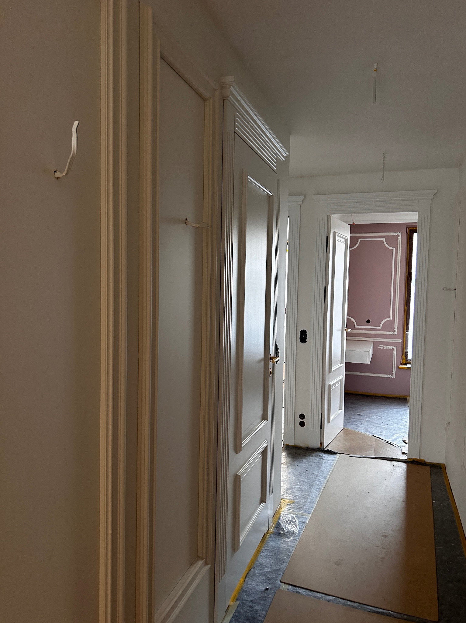 Korytarz w trakcie wykańczania: białe ściany z listwami, zabezpieczona podłoga, widoczne drzwi do pokoju z dekoracyjną ścianą w odcieniach różu i bieli.