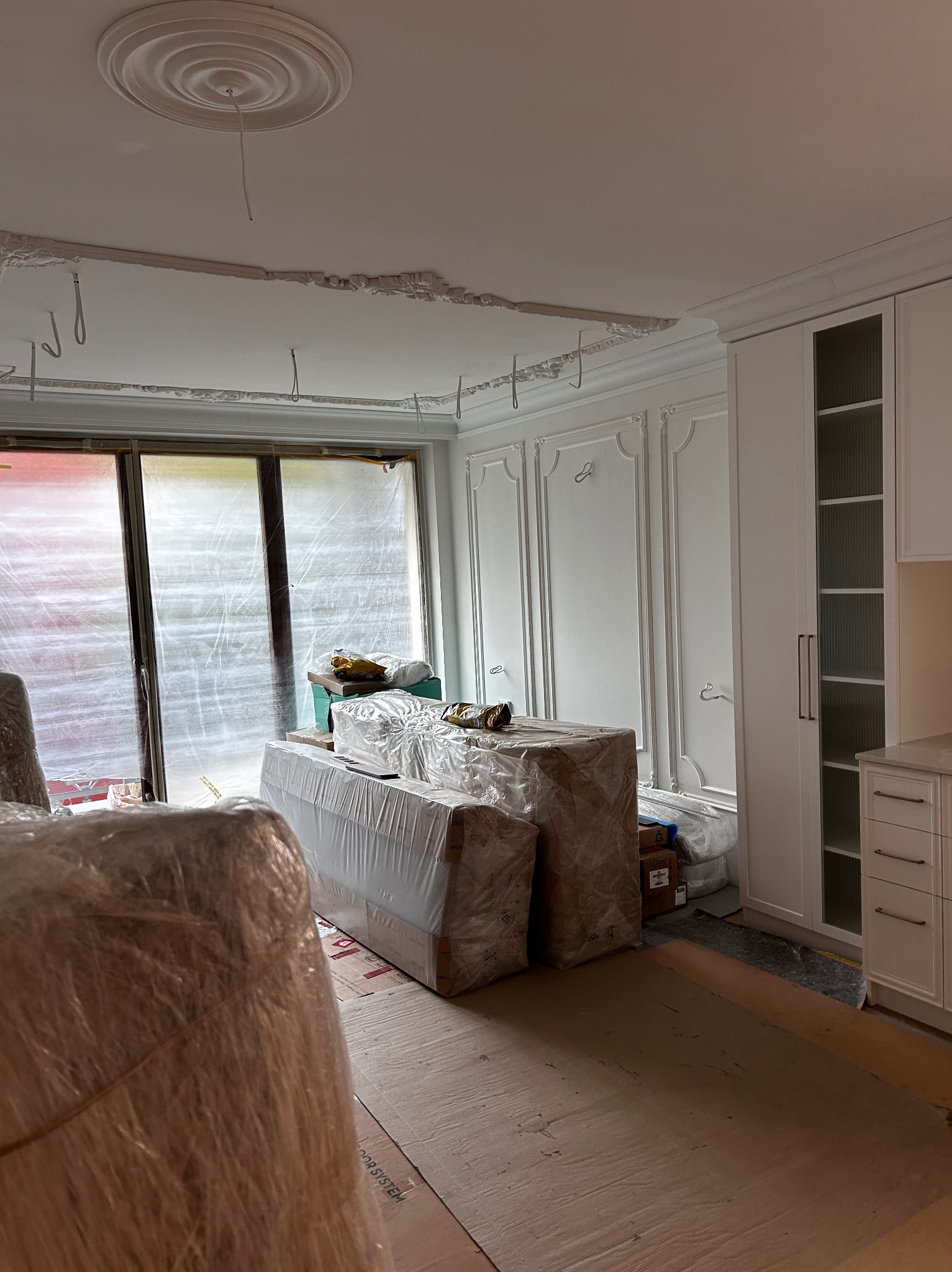 Wnętrze w trakcie wykończenia: zabezpieczone folią meble, okna osłonięte, widoczne elementy dekoracyjne sufitu i ścian, zabudowa meblowa w stylu klasycznym.