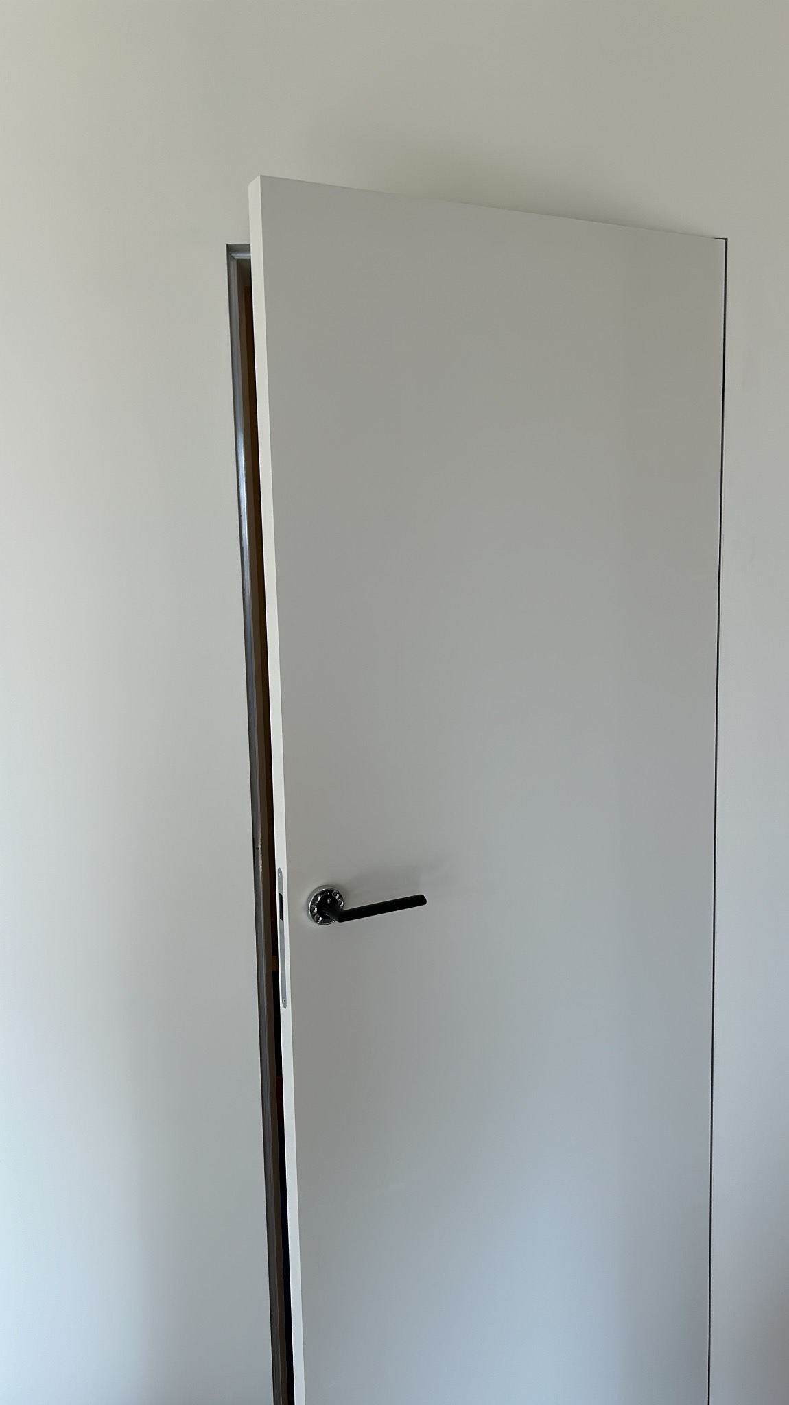 Minimalistyczne, białe drzwi wewnętrzne uchylone, z czarną klamką na tle jasnej ściany. Widoczna ościeżnica i fragment pomieszczenia za drzwiami.