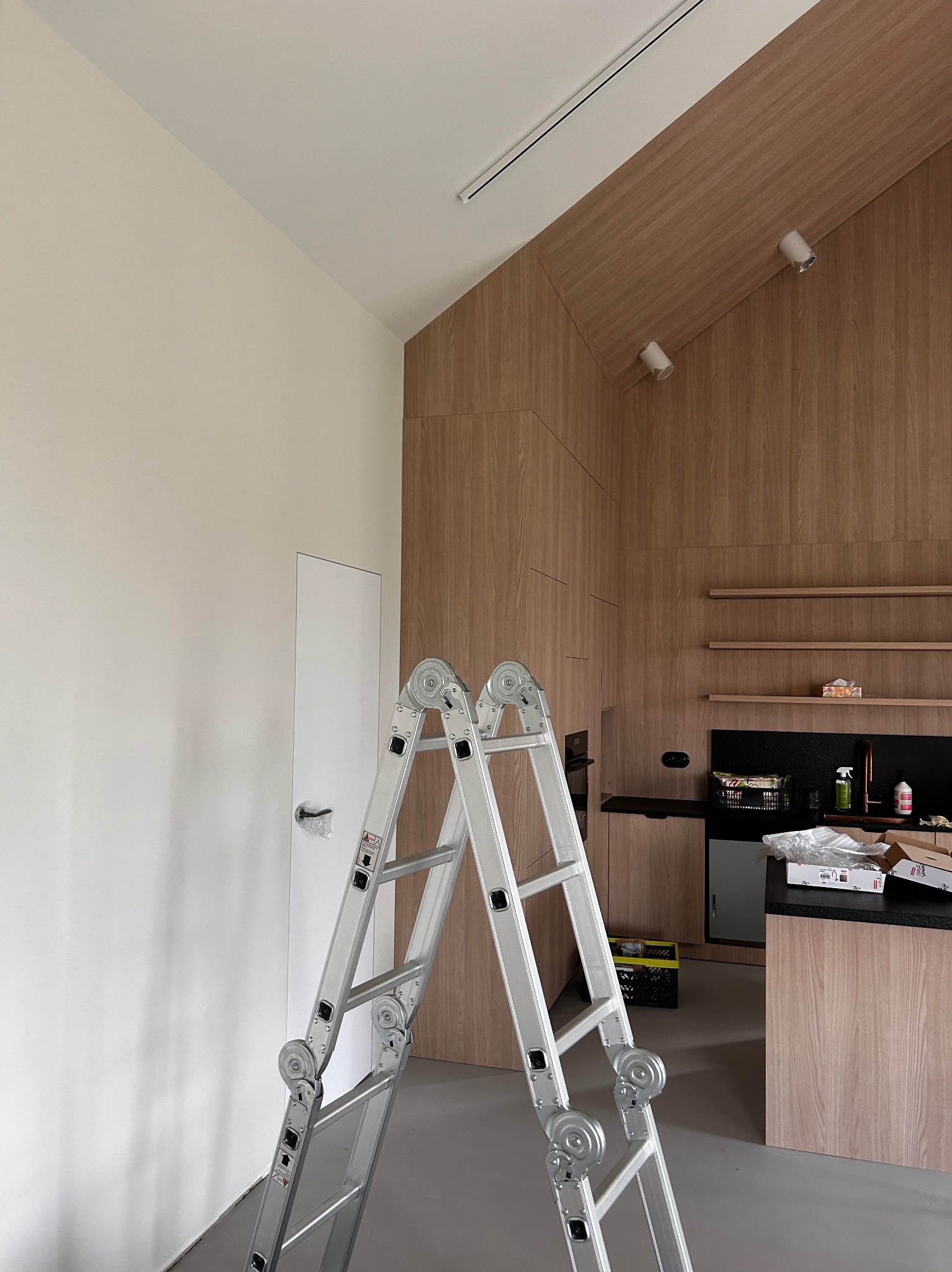 Wnętrze w trakcie remontu: biała ściana, aluminiowa drabina rozstawiona na szarym podłożu, w tle zabudowa kuchenna z drewnianymi frontami i czarnym blatem.
