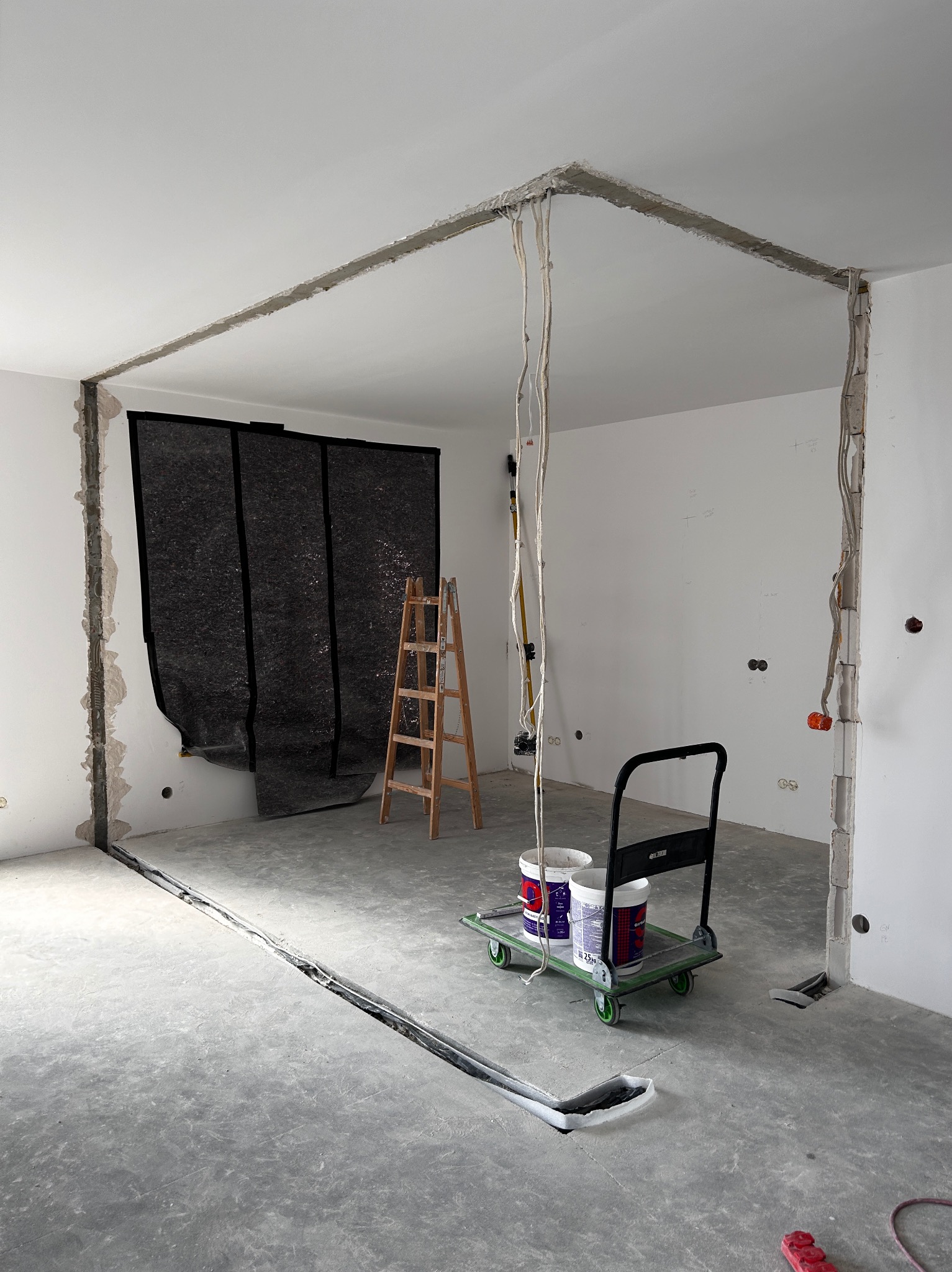 Wnętrze w trakcie prac remontowych: wycięte bruzdy w ścianach i podłodze, drabina, wózek z farbami. Przygotowanie do prac wykończeniowych, widoczne instalacje.