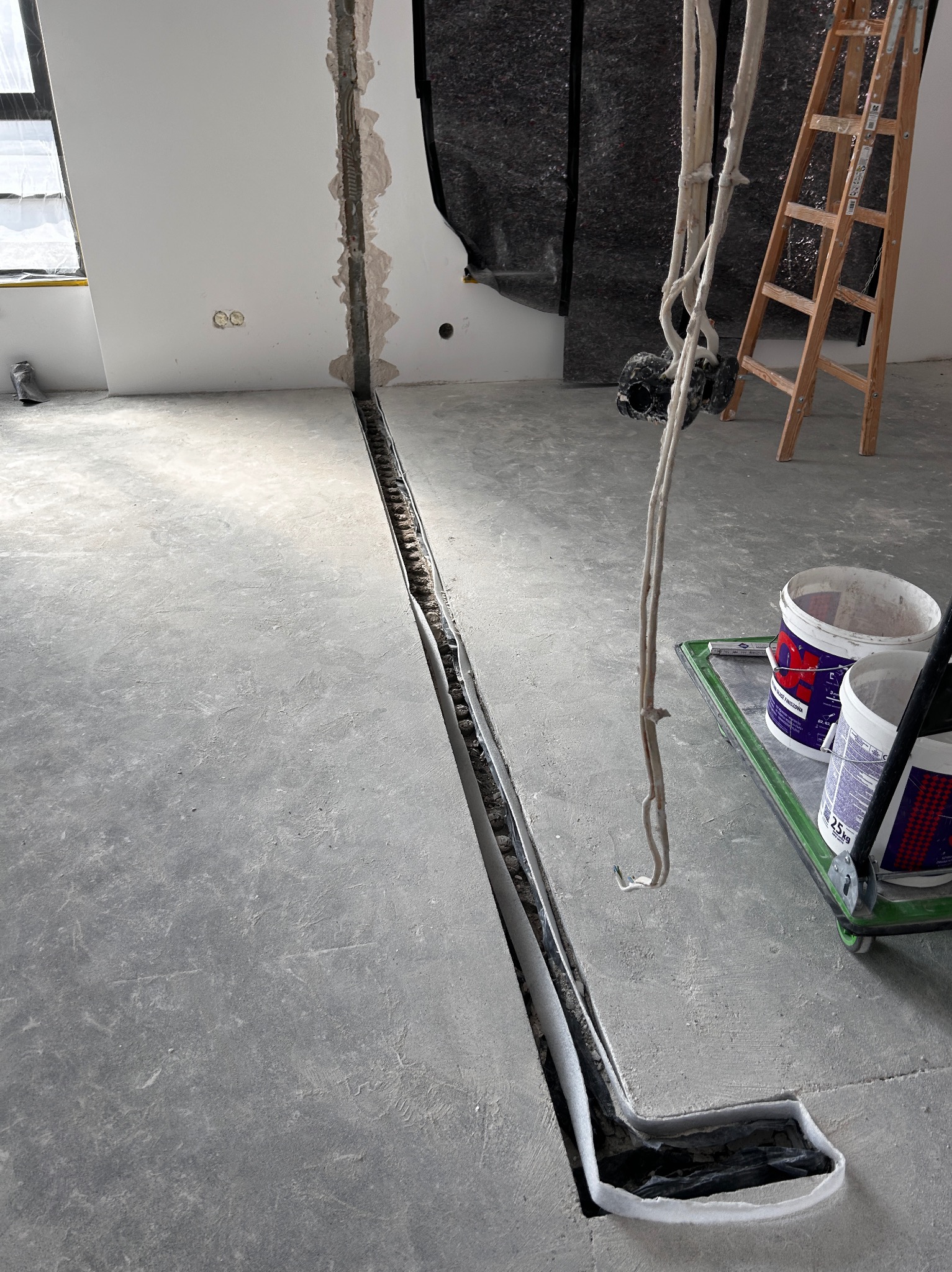 Przygotowanie podłoża: wycięty kanał w posadzce, zabezpieczony folią, widoczne kable, drabina i wiadra z farbą na wózku. Etap prac wykończeniowych.