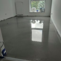 Tomasz Wiatrowski - Wylana, mokra, samopoziomująca wylewka podłogowa w jasnym pomieszczeniu z oknem, widoczne narzędzia i materiały budowlane na parapecie.