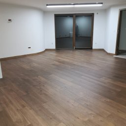 Tomasz Wiatrowski - Ułożone panele podłogowe w pustym pomieszczeniu z białymi ścianami, widoczne listwy przypodłogowe i duże okno balkonowe.