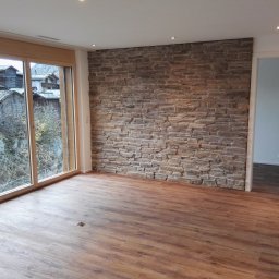 Tomasz Wiatrowski - Wnętrze pokoju z drewnopodobną podłogą, kamienną ścianą i widokiem na górską wioskę przez duże okno.