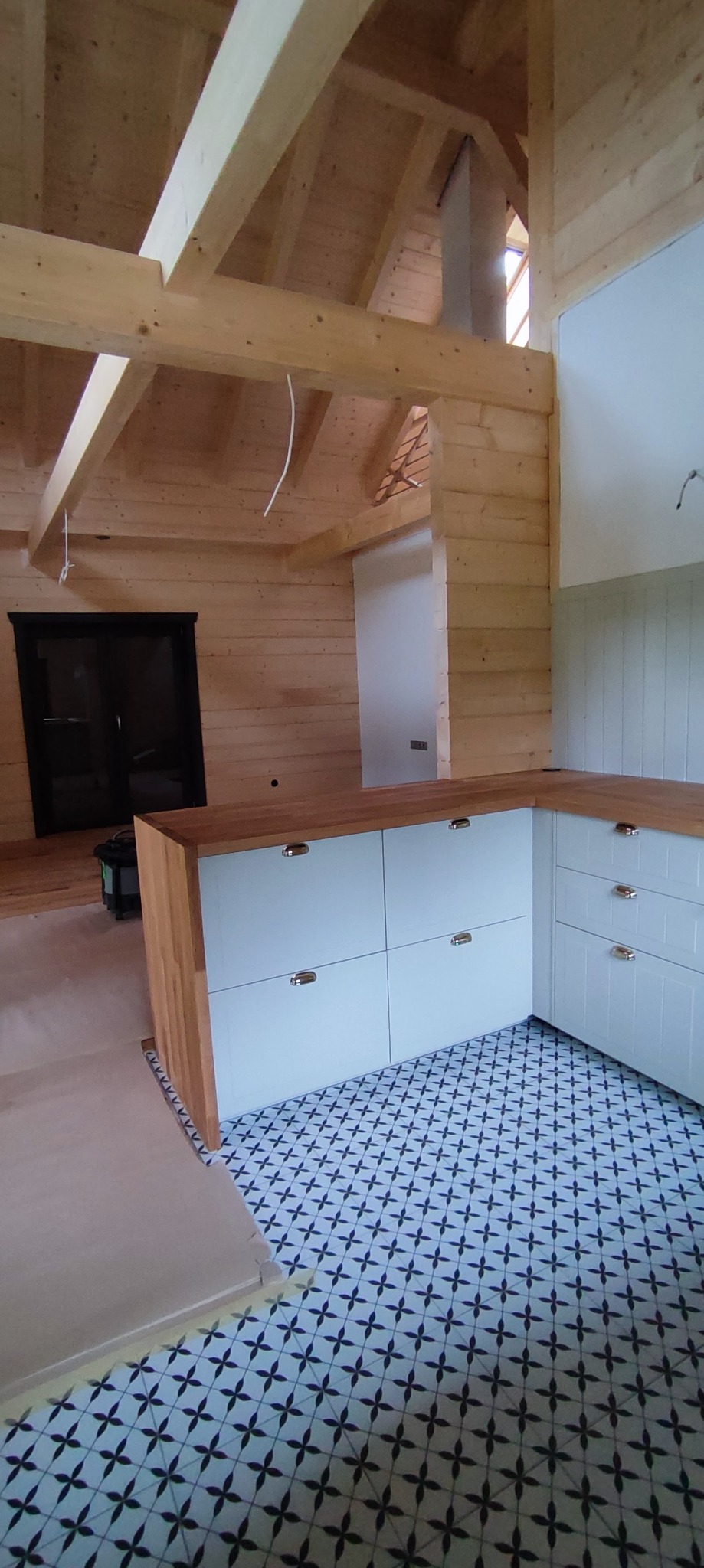 Wnętrze drewnianego domu z jasnymi szafkami kuchennymi z drewnianymi blatami i wzorzystą podłogą, widoczne surowe drewniane belki stropowe i instalacje.