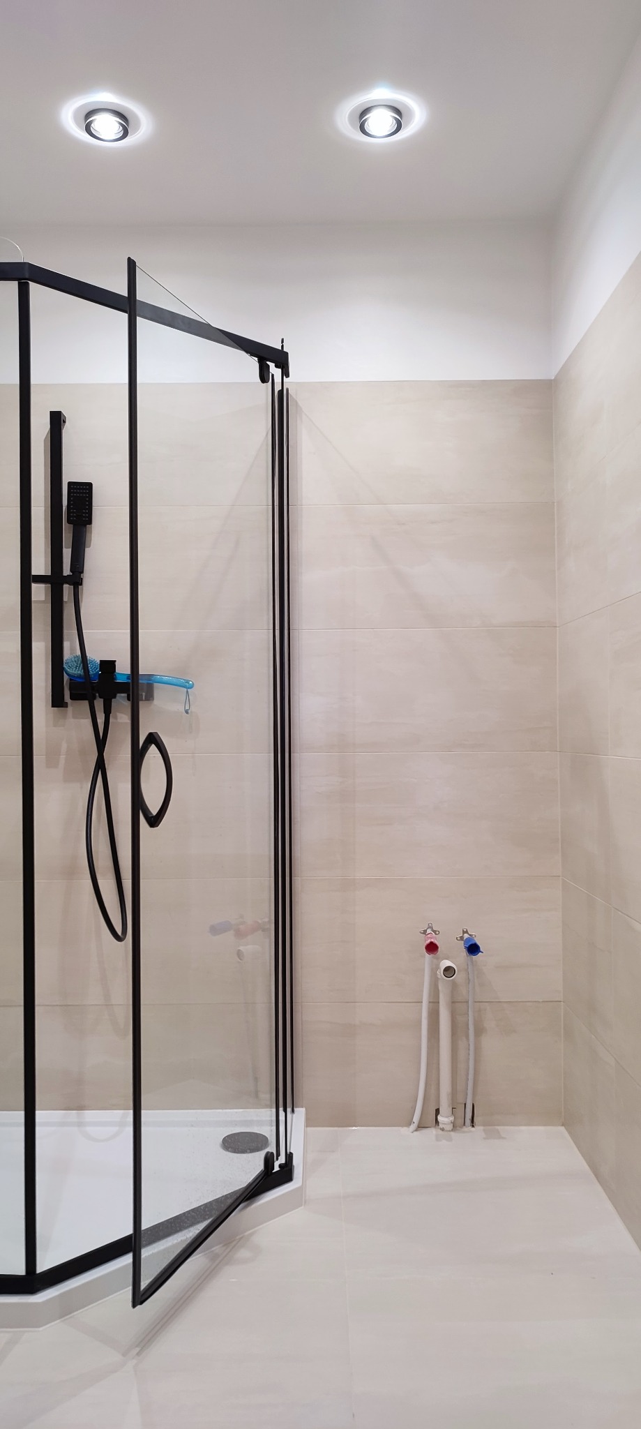 Nowoczesna kabina prysznicowa z czarnymi profilami, jasne płytki ceramiczne na ścianach i podłodze, widoczne przyłącza wodne z czerwonym i niebieskim zaworem.