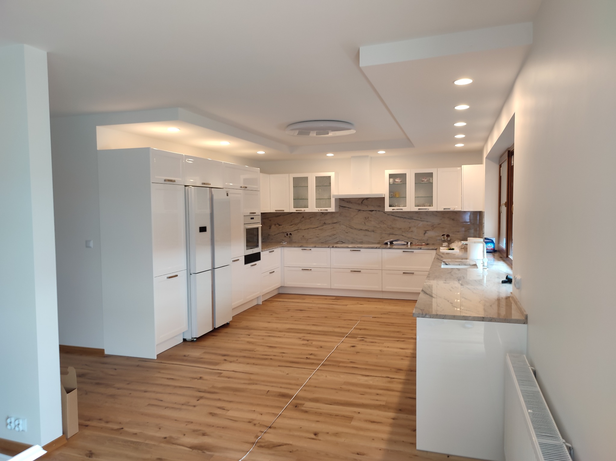 Jasna kuchnia z białymi, błyszczącymi szafkami, marmurowym blatem i drewnianą podłogą, oświetlona punktowymi lampami sufitowymi.