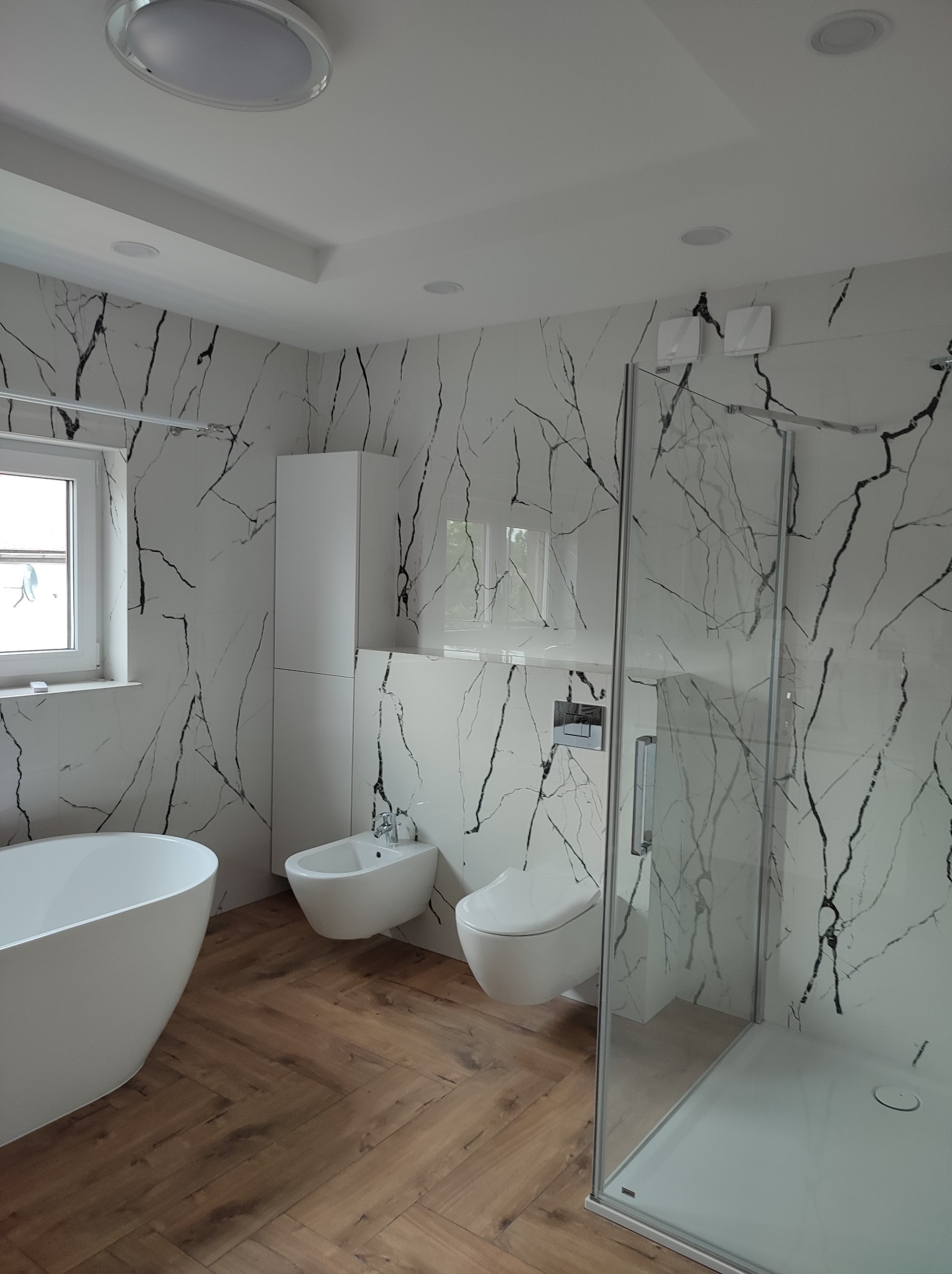 Nowoczesna łazienka z białą armaturą, płytkami imitującymi marmur z czarnym żyłowaniem i drewnopodobną podłogą ułożoną w jodełkę, oświetlona światłem punktowym.