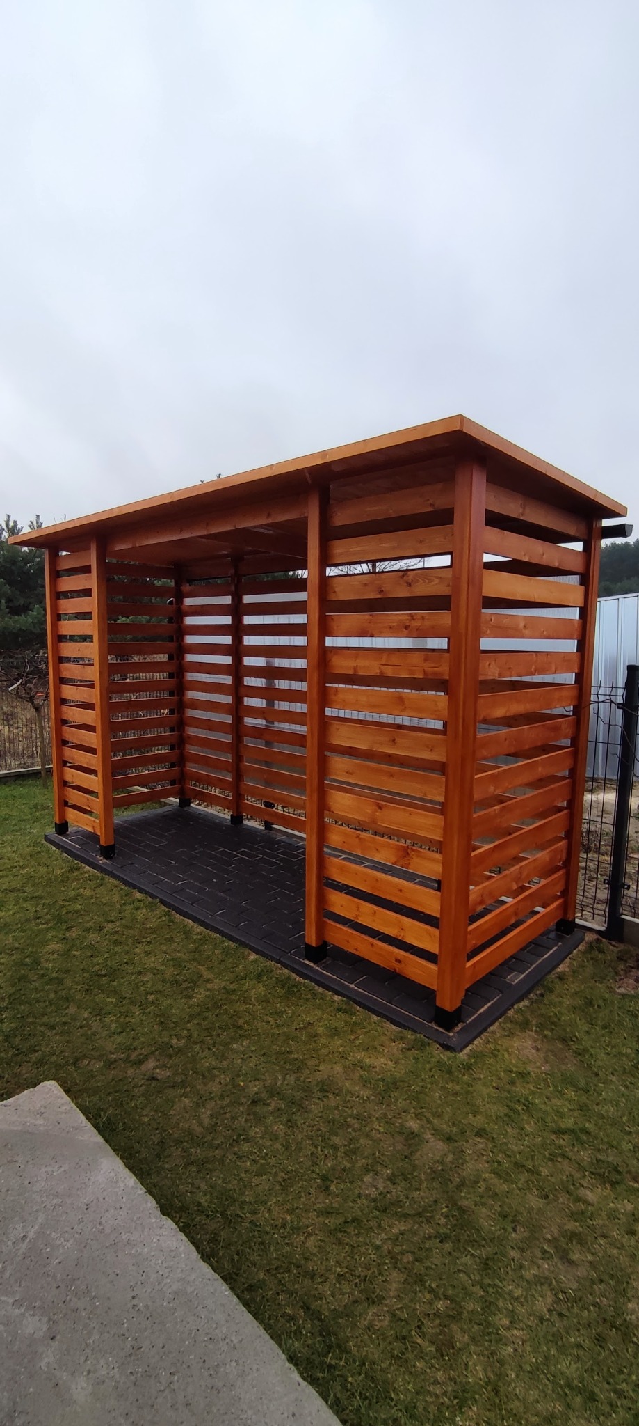 Nowoczesna, drewniana altana z poziomymi listwami, stojąca na czarnym, brukowanym podłożu na trawniku.
