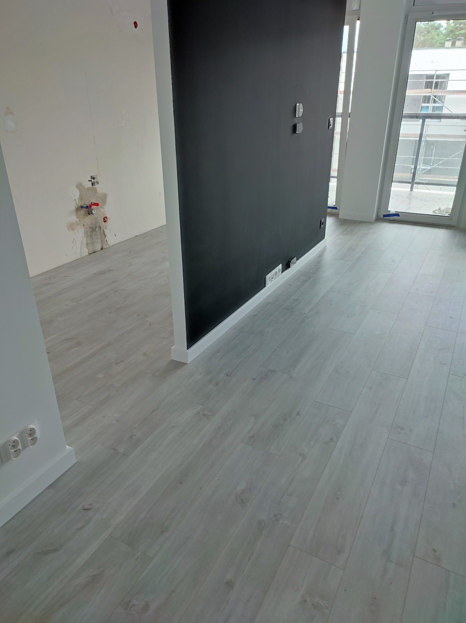 Jasnoszare panele podłogowe ułożone w nowym mieszkaniu, widoczna czarna ściana działowa z gniazdkami, fragment ściany z instalacją hydrauliczną przed wykończeniem.