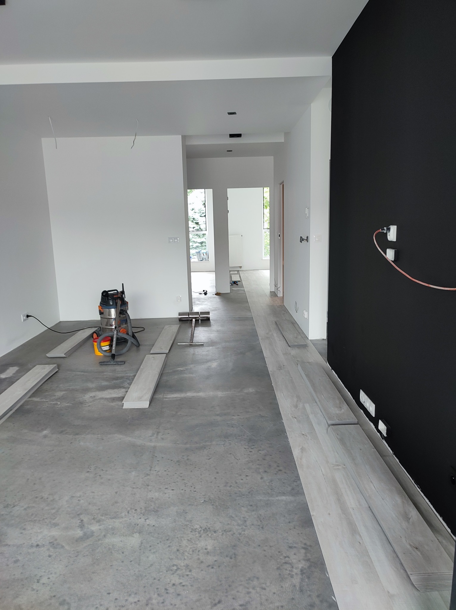 Przygotowanie do układania jasnych paneli podłogowych w przestronnym, częściowo wykończonym wnętrzu z czarną ścianą akcentującą, widoczny odkurzacz przemysłowy i narzędzia pomiarowe.